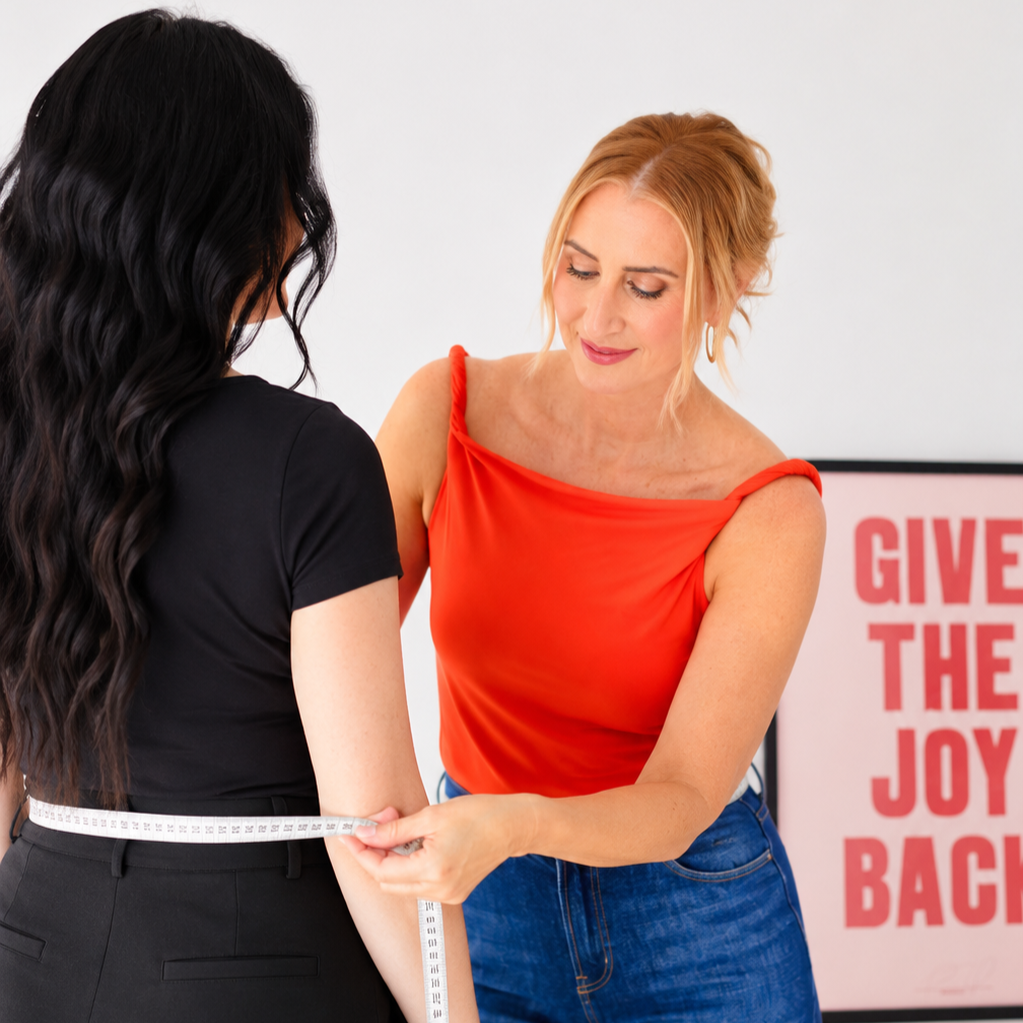A woman in a red top is measuring the arm of another woman in black with a measuring tape, in front of a pink sign that says "GIVE THE JOY BACK."