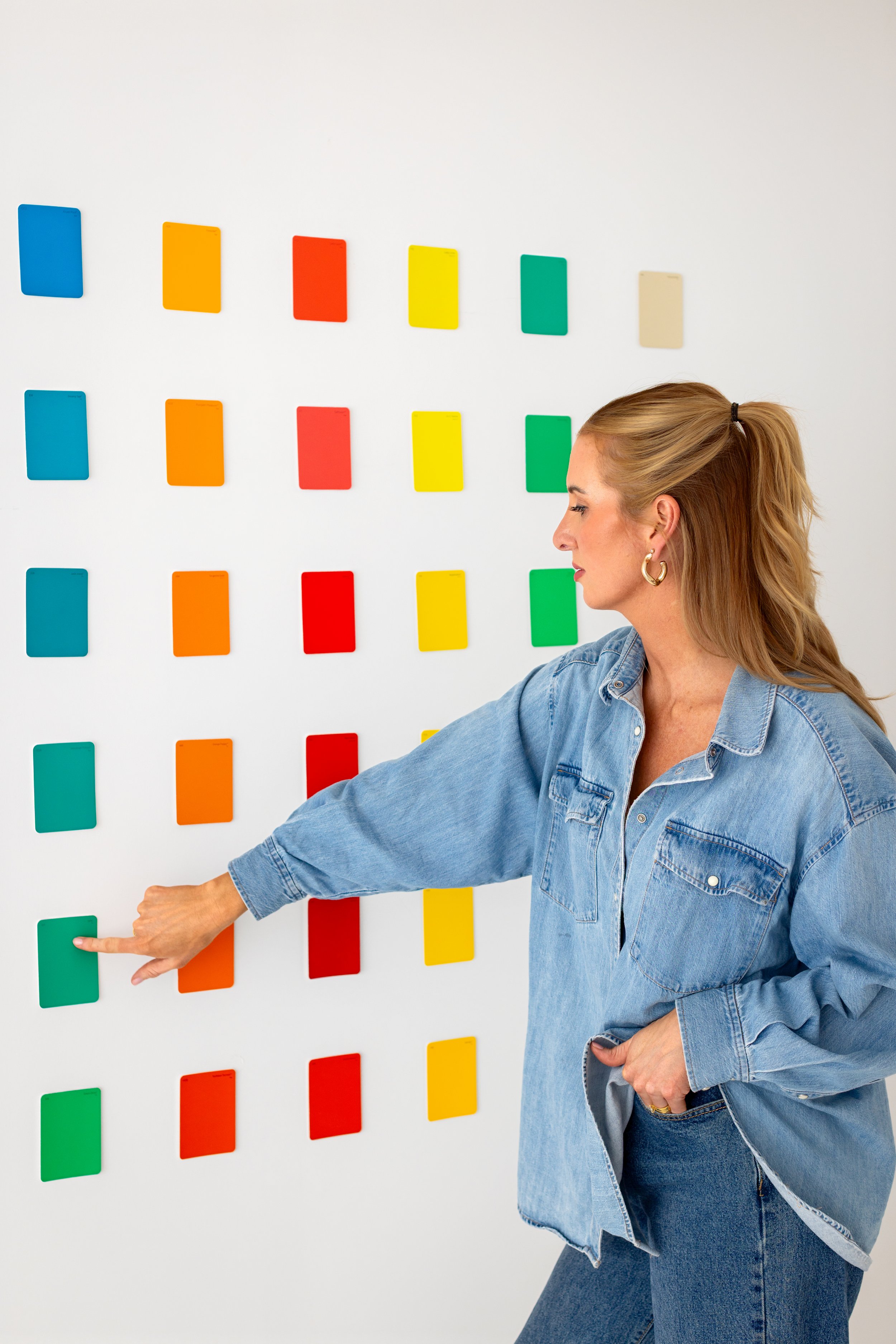 A woman in a denim jacket and jeans points at a colorful wall of rectangular cards, including blue, yellow, red, and green, arranged in a grid pattern.