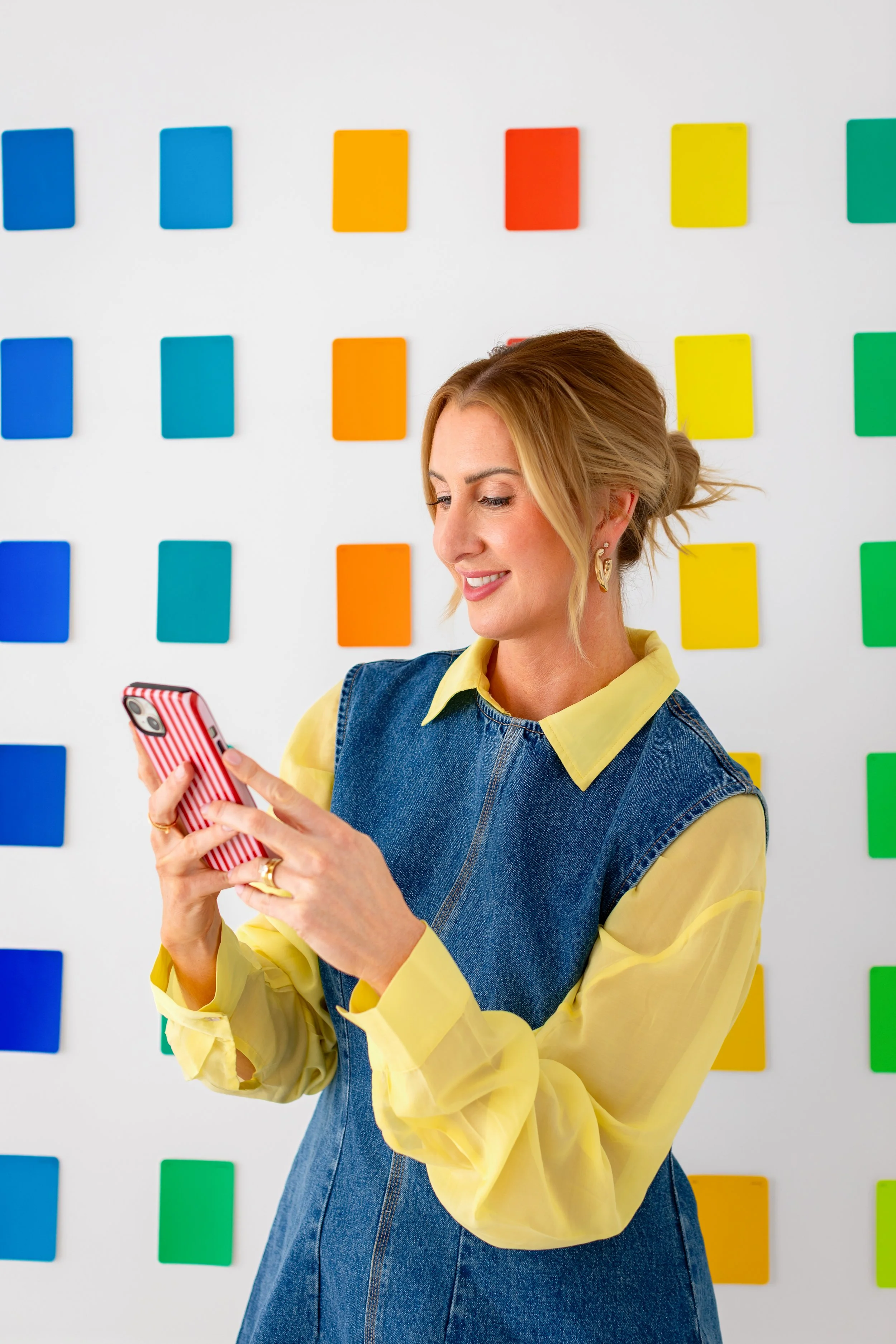 A woman with blonde hair styled in a messy bun, wearing a yellow blouse and denim vest, smiling while looking at her phone against a white wall with colorful cards arranged in rows.