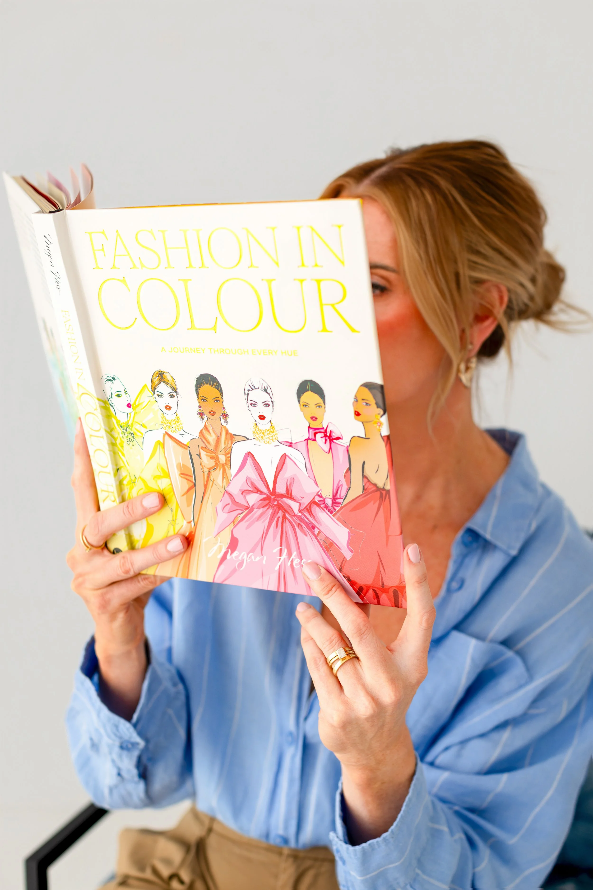 A woman with blonde hair, wearing a blue button-up shirt, with her face partially obscured by a book titled 'Fashion in Colour' she is holding. The book features illustrations of women dressed fashionably.