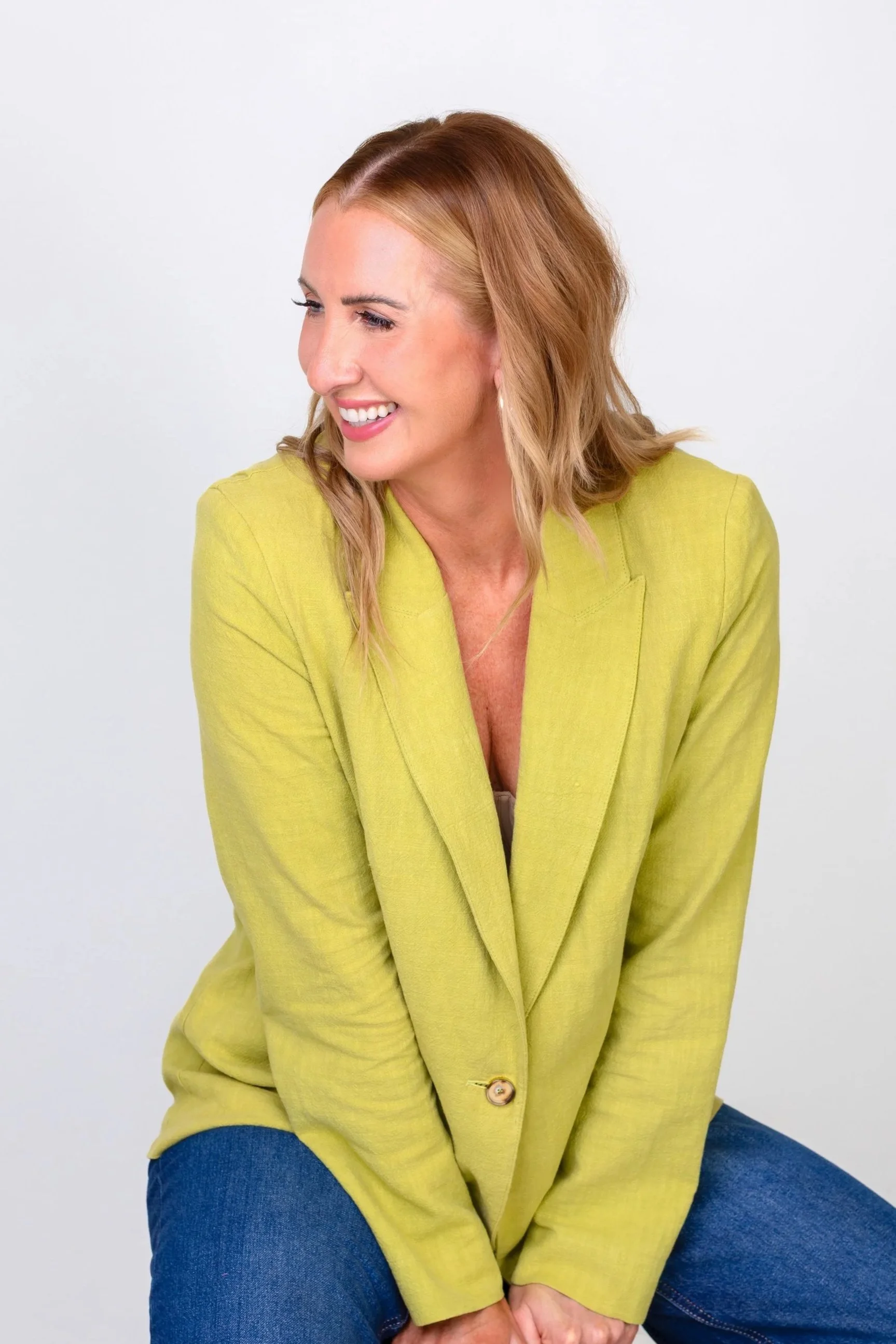 A woman with shoulder-length blonde hair, wearing a lime green blazer with a button, sitting against a plain white background. She is smiling slightly and looking to her left.
