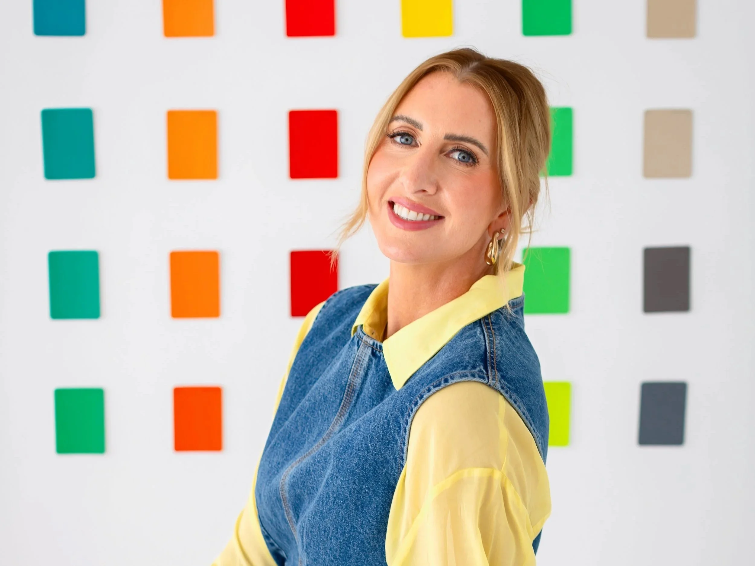 A smiling woman with blonde hair and blue eyes, wearing a yellow shirt and denim jacket, stands in front of a white wall decorated with colorful rectangular tiles.
