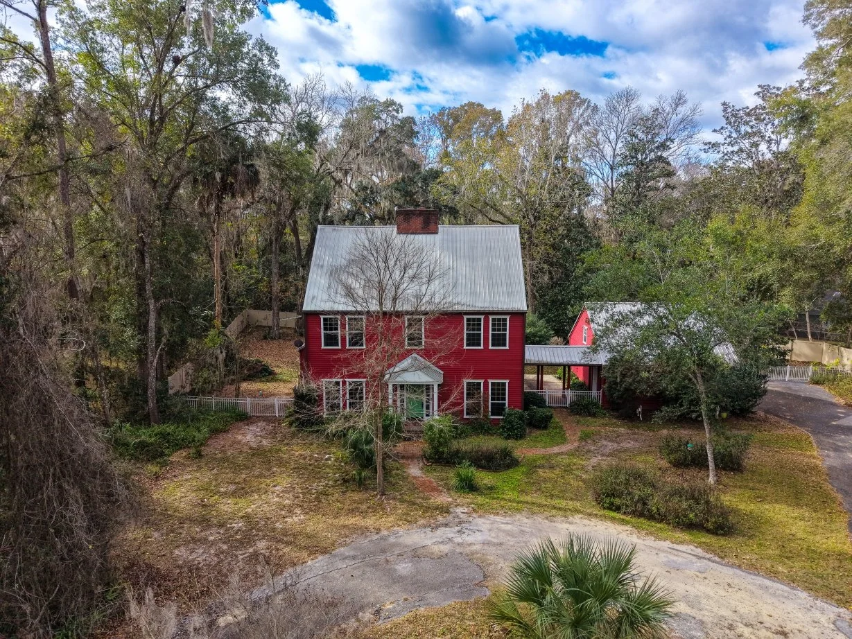 A red two-story house with a white metal roof, surrounded by trees and a yard, with a driveway on the right side leading to a small outbuilding.