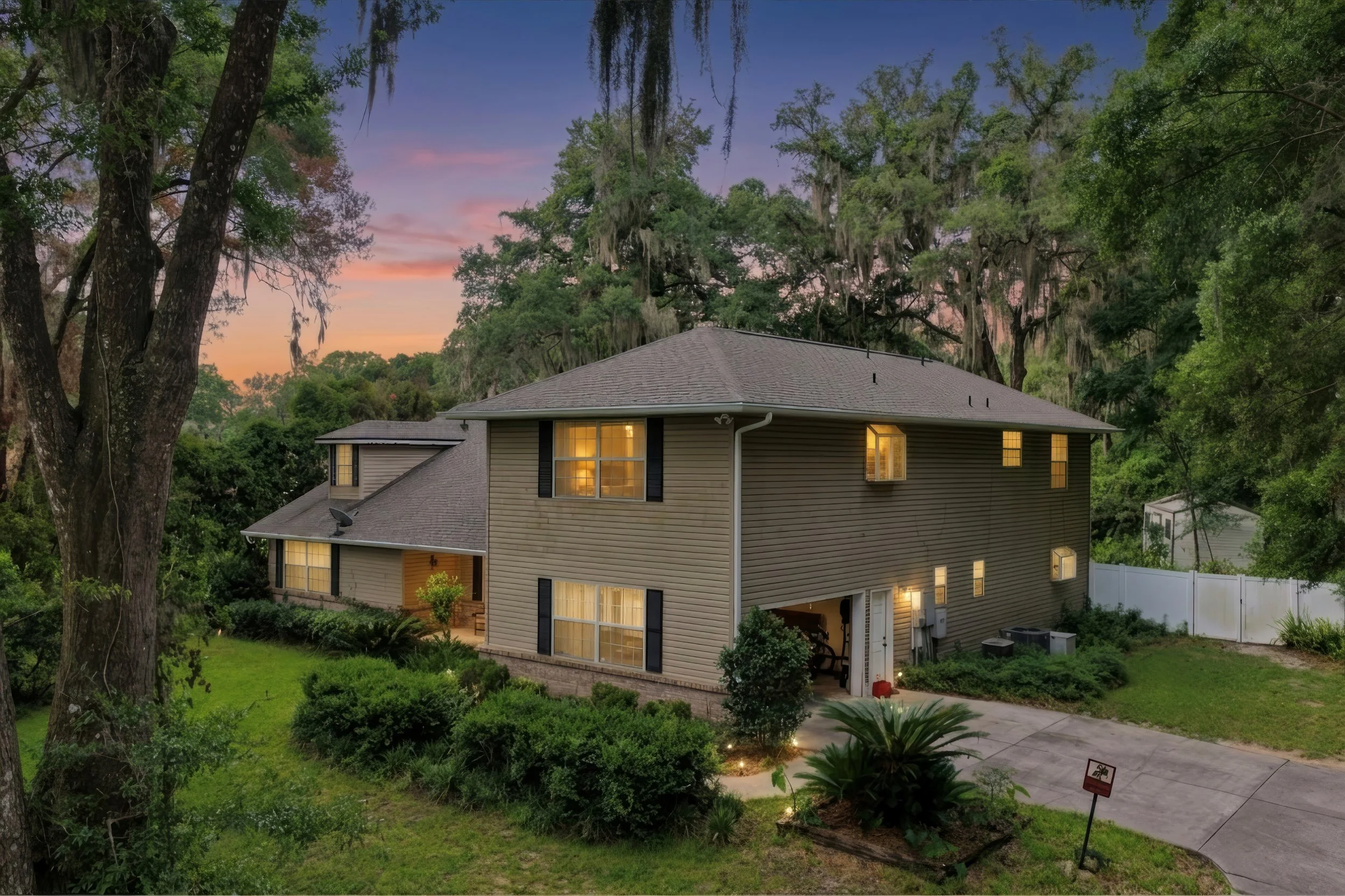 A two-story house with beige siding and a dark gray roof surrounded by lush green trees and a hillside, with lights on inside the house at dusk.