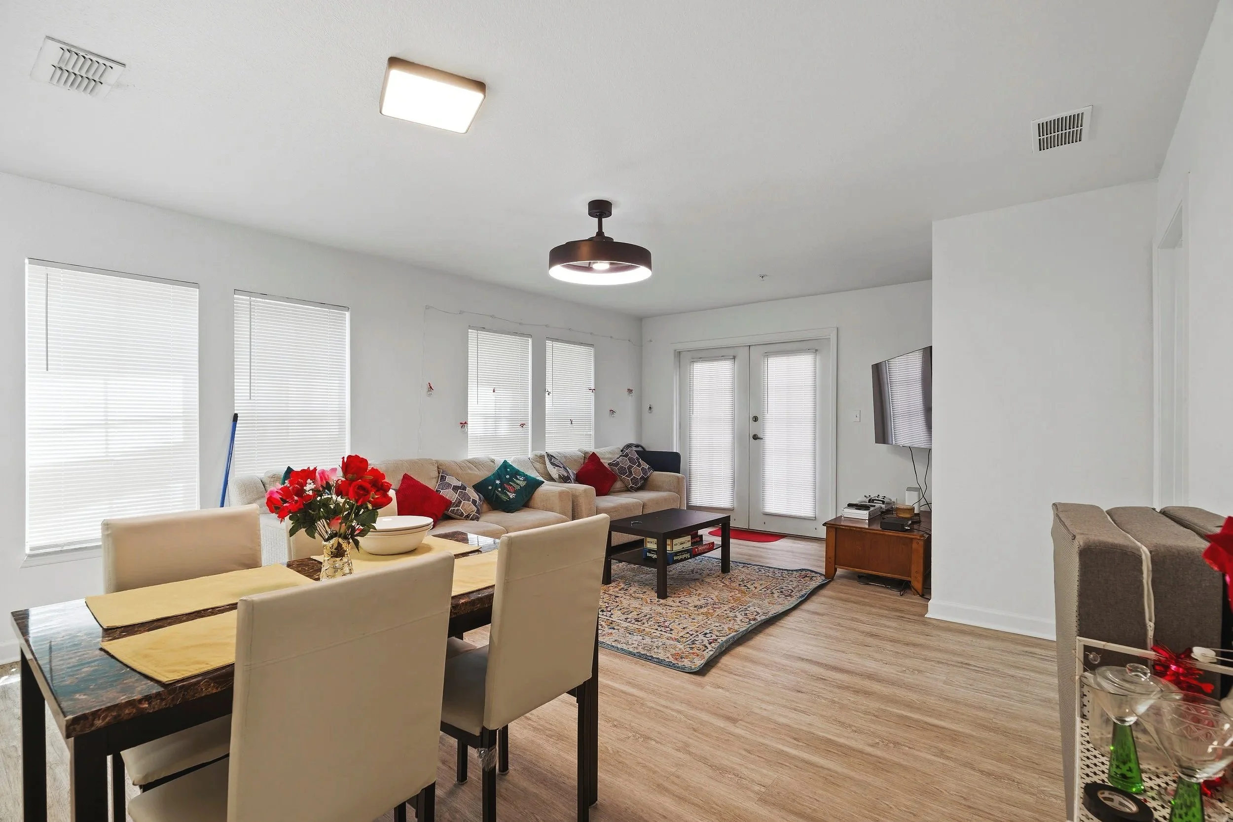 Living room with a dining table, beige sofa with colorful pillows, coffee table, and television, illuminated by natural light through multiple windows with blinds, with holiday decorations visible.
