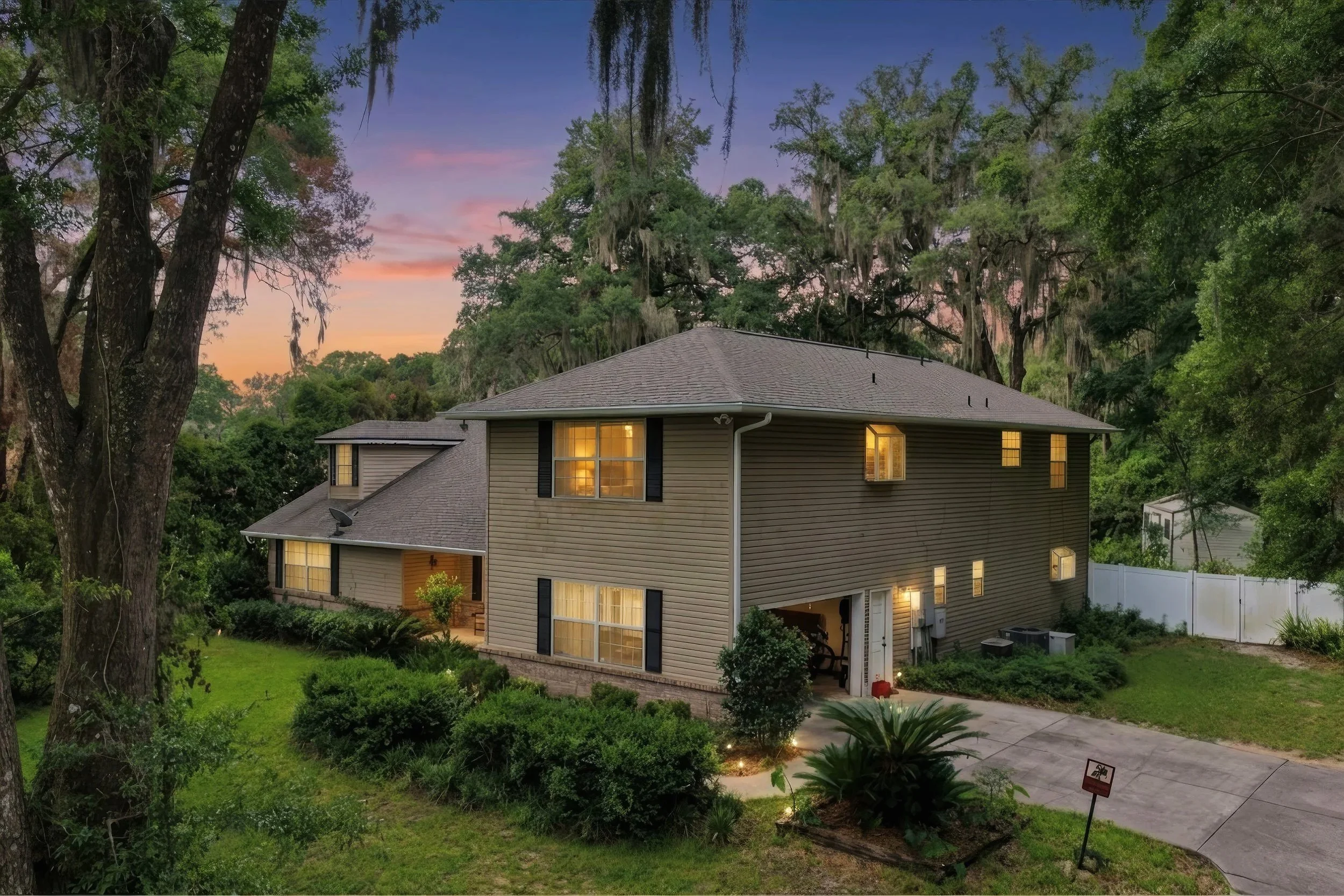 A multi-story house with beige siding and black shutters surrounded by lush green trees at dusk, with lights on inside and a sunset sky visible.