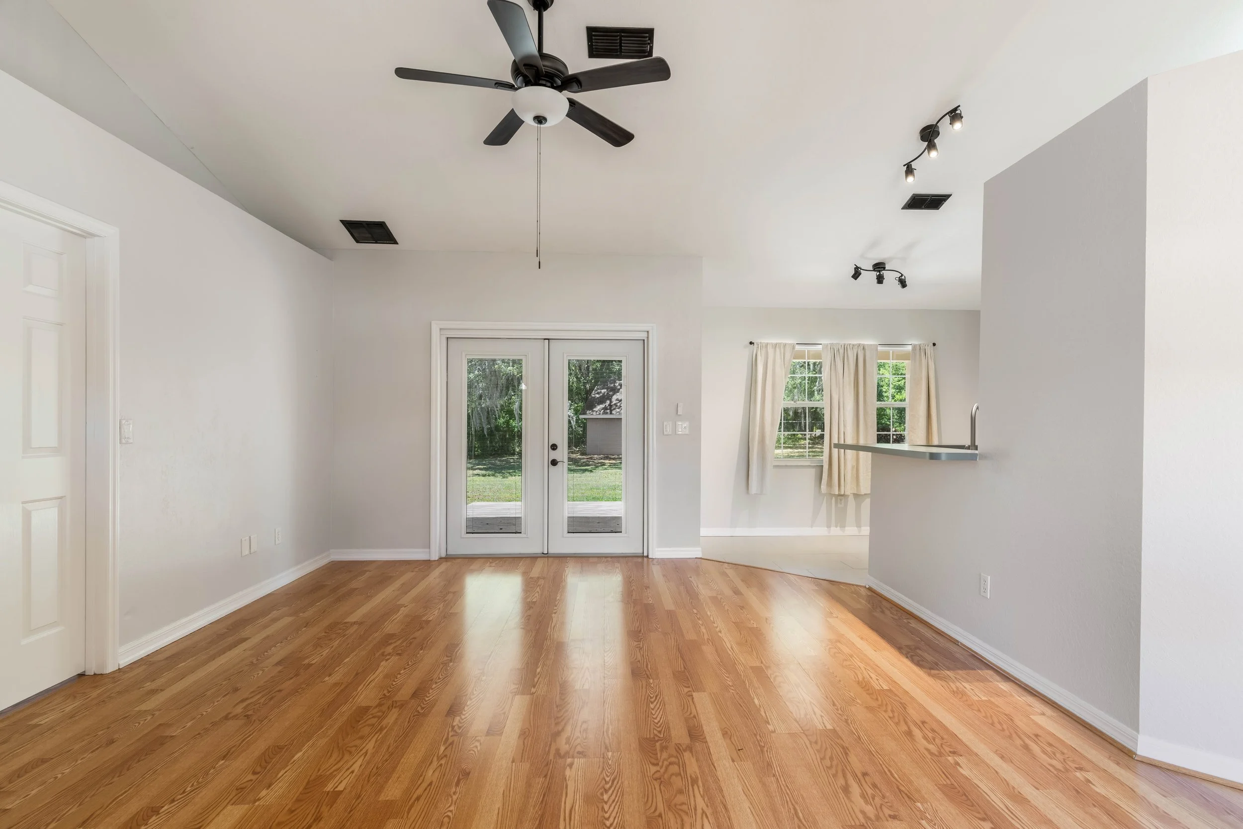 Empty living room with hardwood flooring, white walls, ceiling fan, ceiling lights, glass sliding door, windows with curtains, and partial view of kitchen counter.