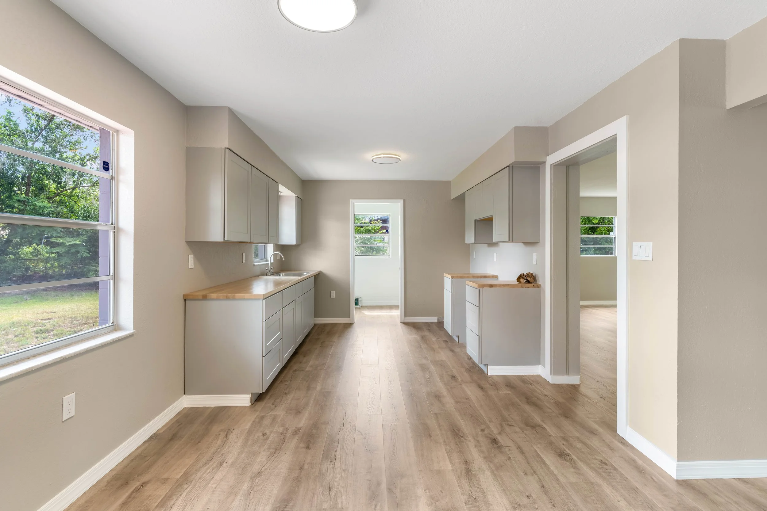 Open-concept kitchen with light gray cabinets, wooden countertops, large windows, and hardwood flooring.