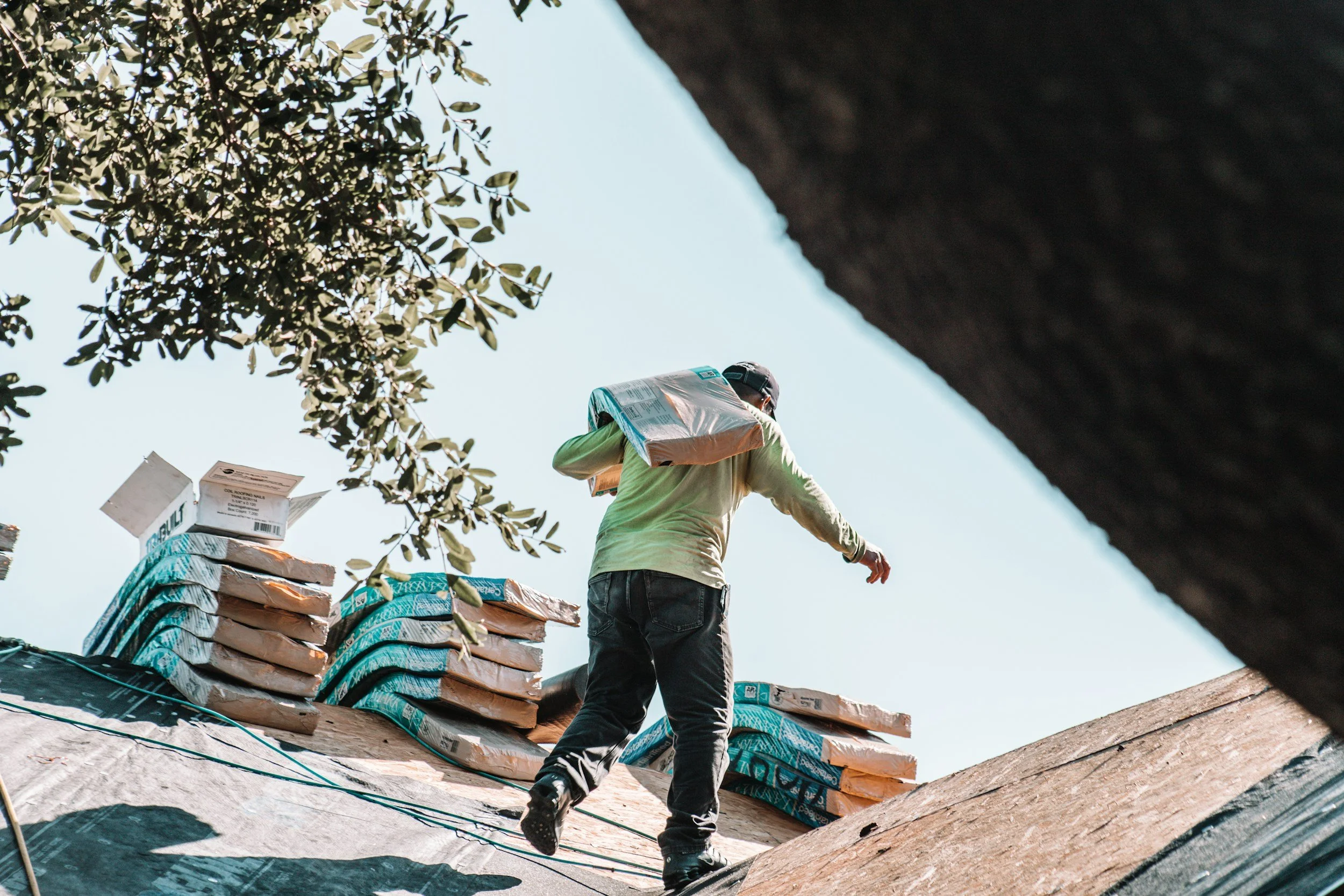 A construction worker climbing a ladder on a roofing site, surrounded by stacks of roofing materials, with a tree in the background and a clear sky.