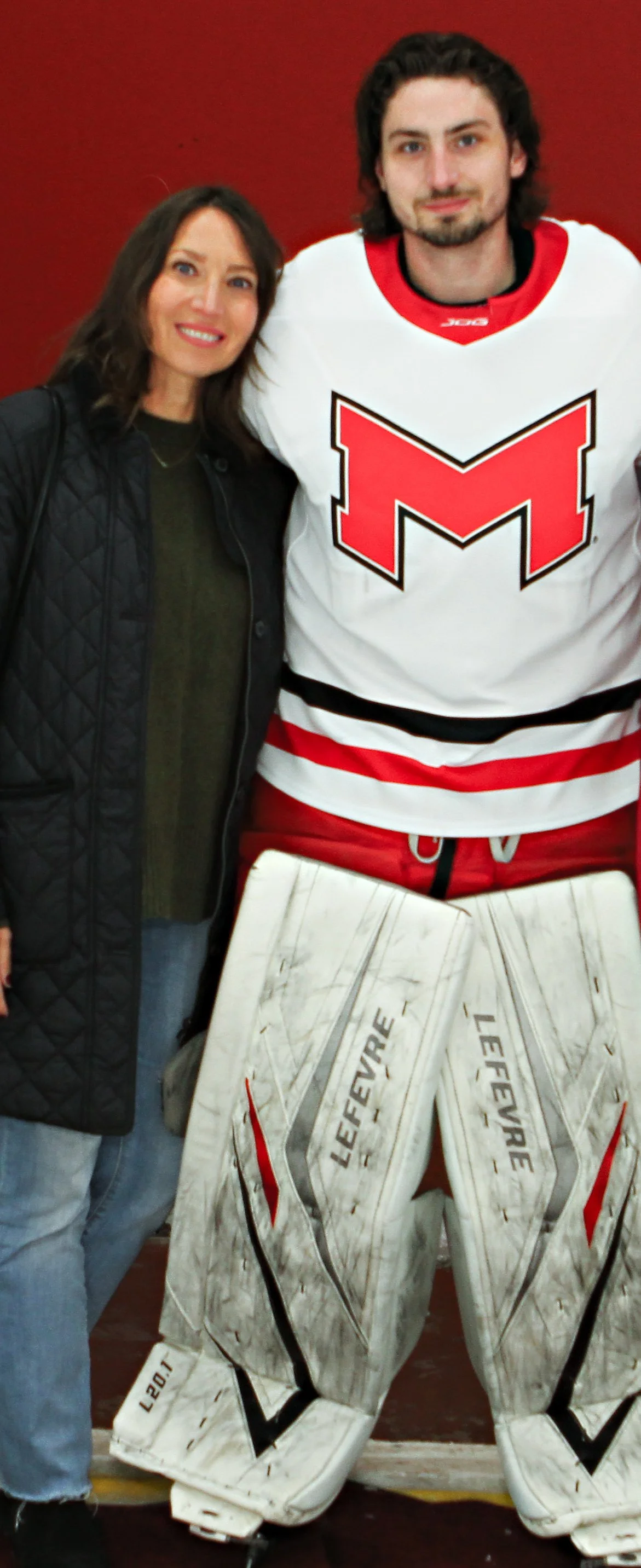 A woman and a hockey player standing together, posing for a photo, with the hockey player wearing a white and red team jersey and goalie pads.