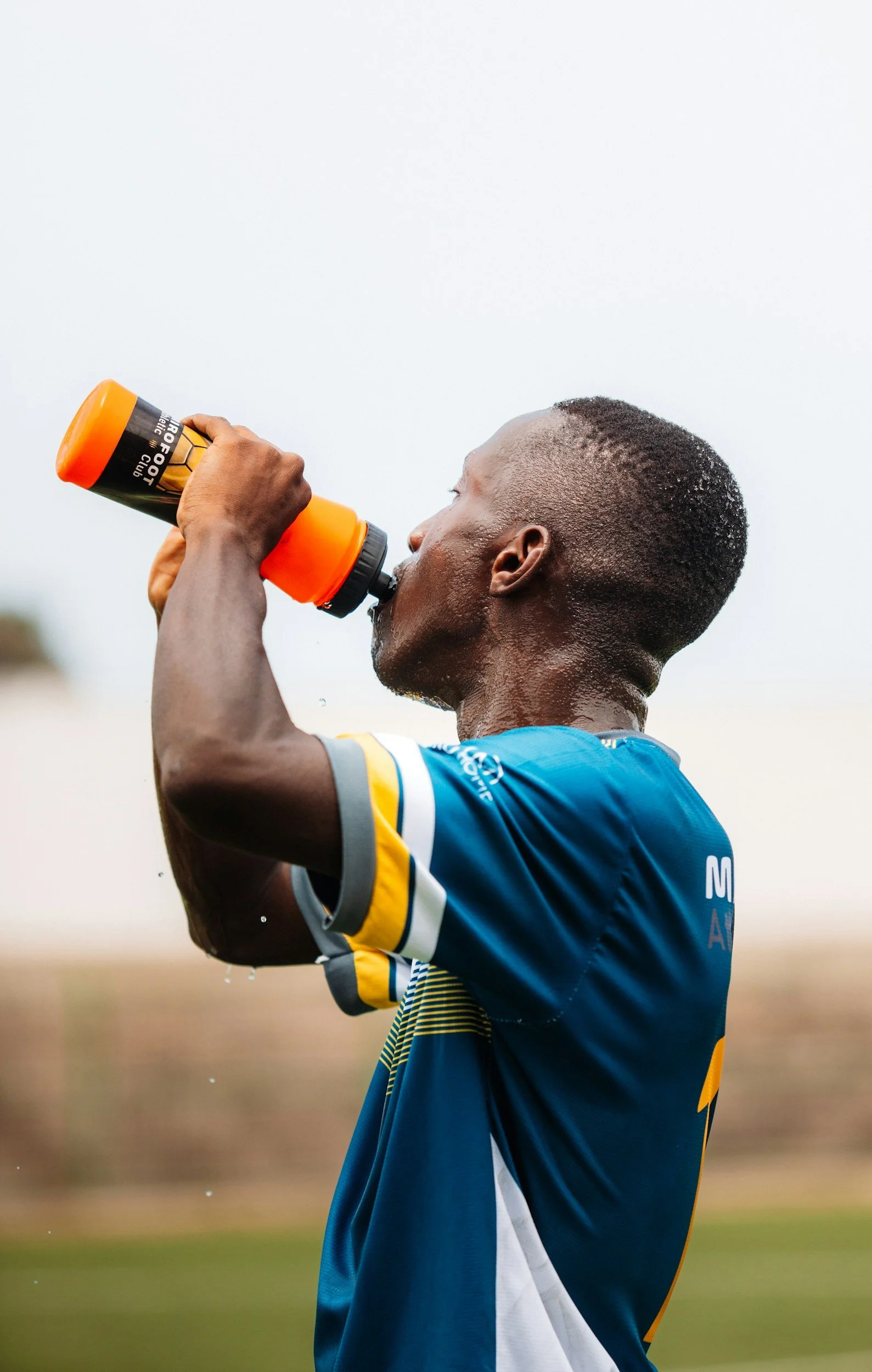 A male athlete in a blue and yellow sports jersey drinking water from an orange bottle during a game or practice outdoors.