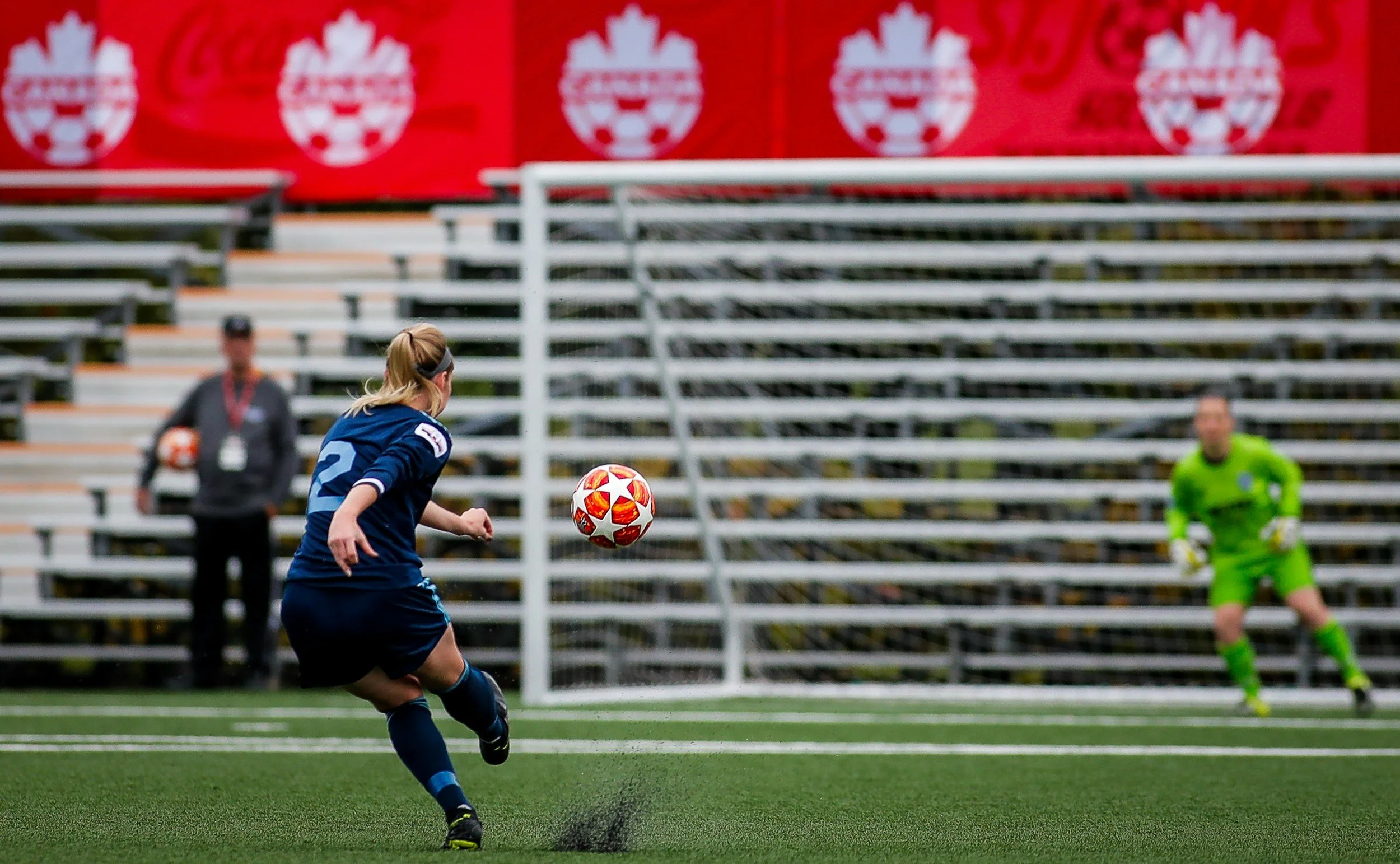 A female soccer player in a blue jersey kicks a soccer ball on a field during a match, with a goalkeeper in green and spectators' bleachers in the background adorned with Canadian flag banners.