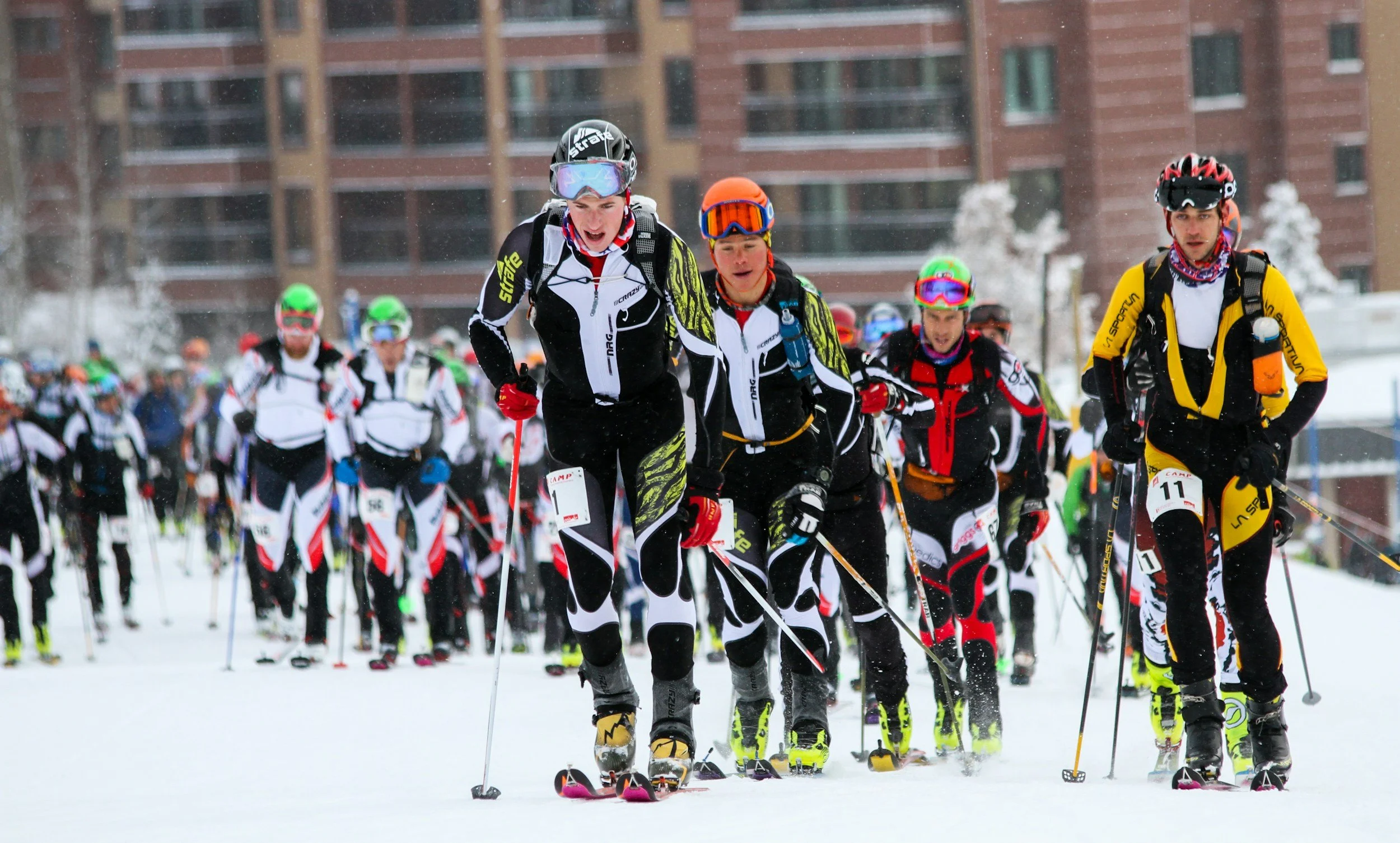 Group of people participating in a cross-country skiing race on a snowy day, with some wearing helmets and goggles, in an urban area with apartment buildings in the background.