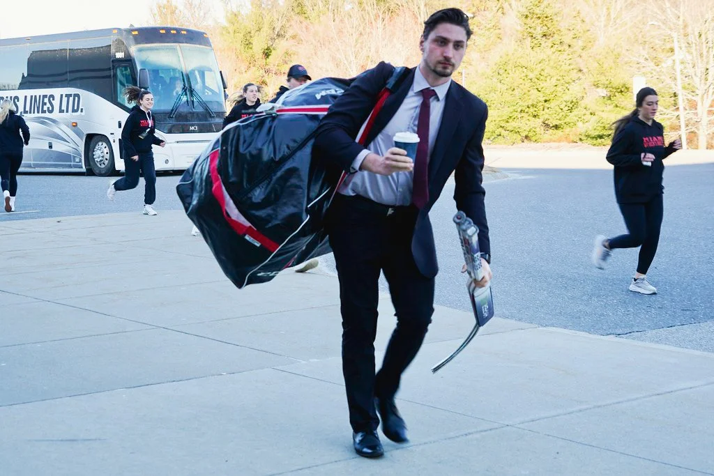 A man in a business suit holding a coffee cup and a hockey stick, walking on a sidewalk with a sports team and a bus in the background.