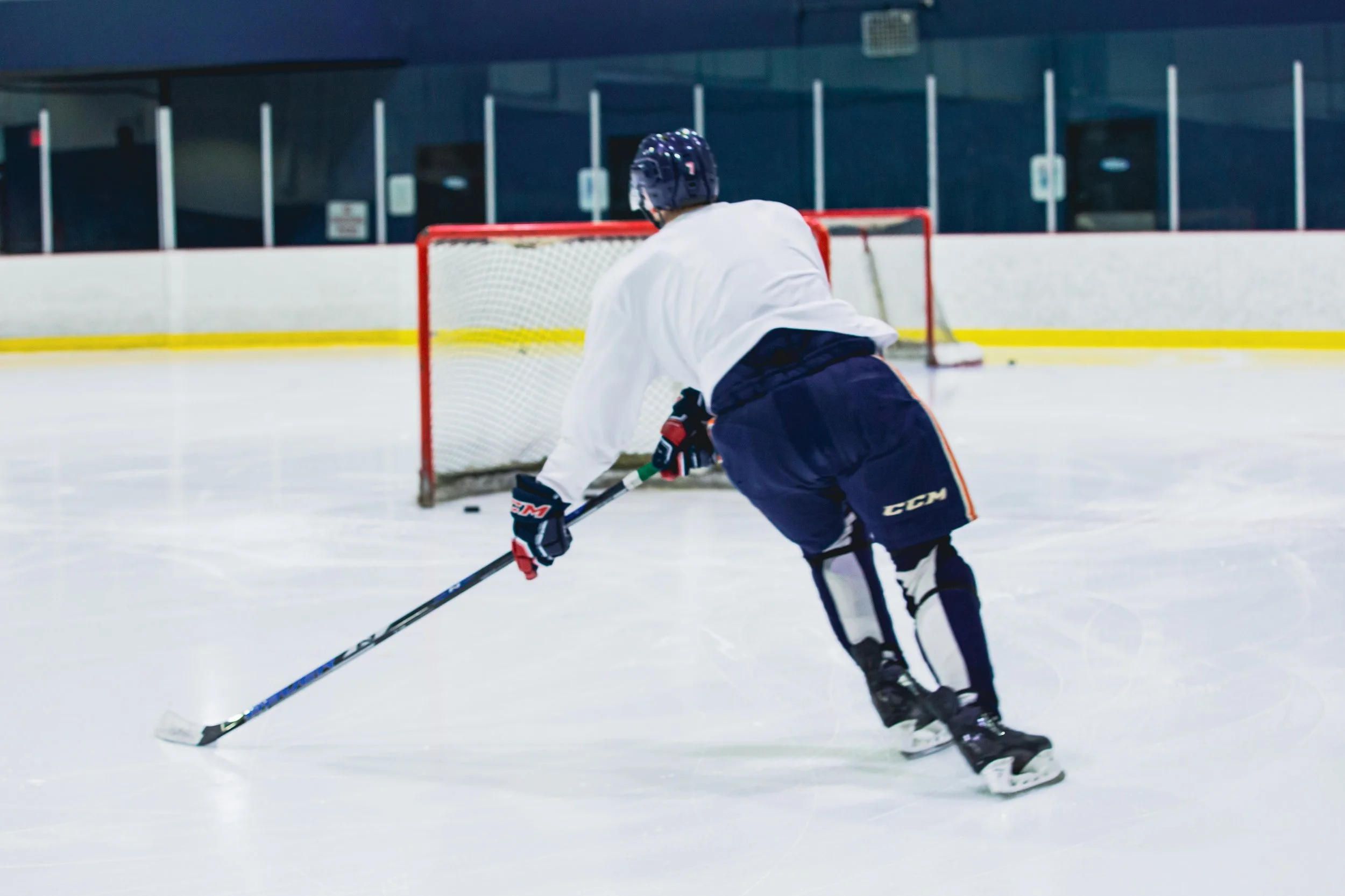 A hockey player in white jersey and black pants, wearing a helmet and gloves, skating on ice rink with a hockey stick, near the goal net.