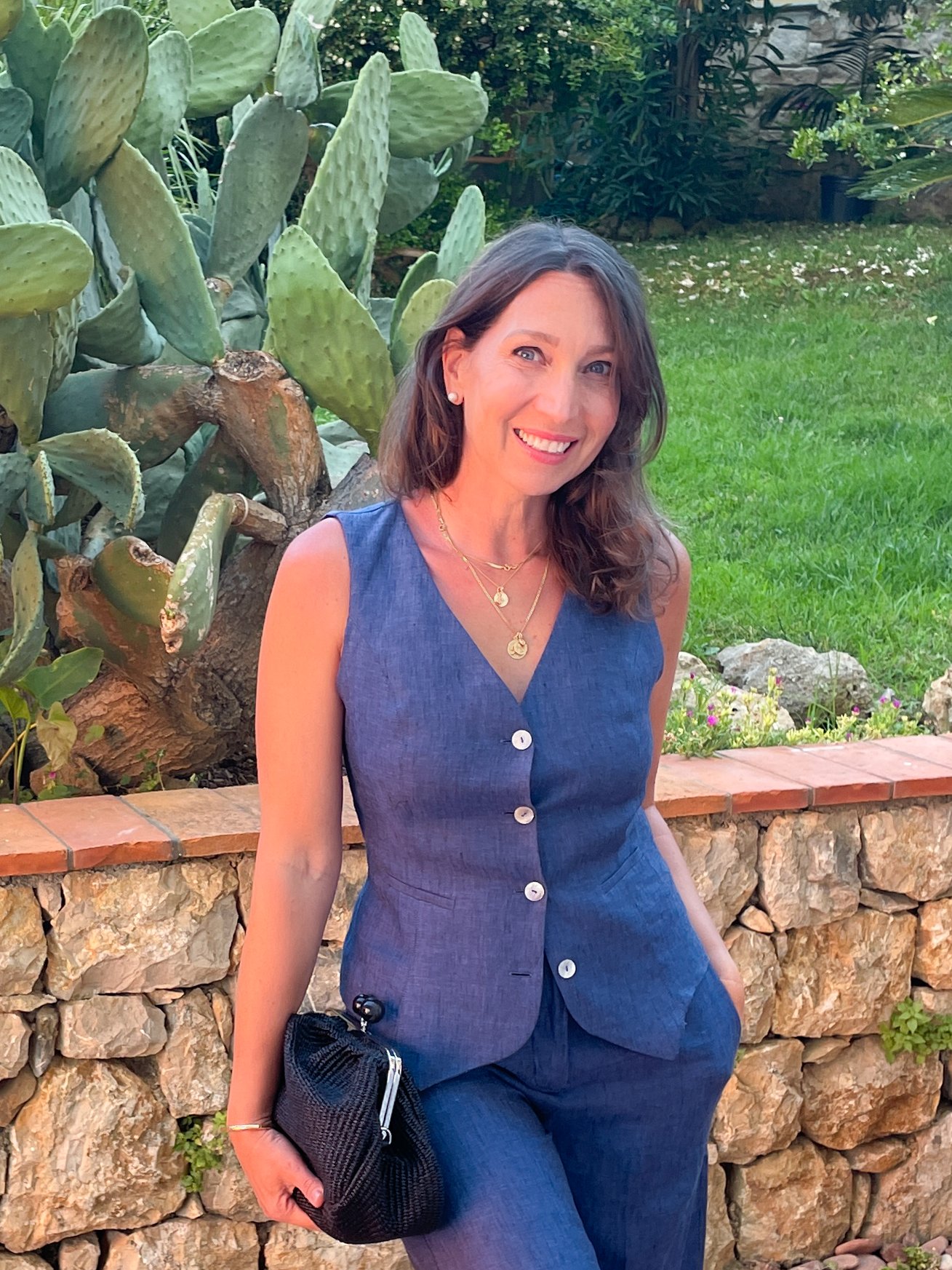 A woman with shoulder-length brown hair and light skin, smiling and wearing a sleeveless blue dress with white buttons, gold jewelry, and holding a black textured clutch, standing outdoors next to a stone wall with a large cactus plant in the background.