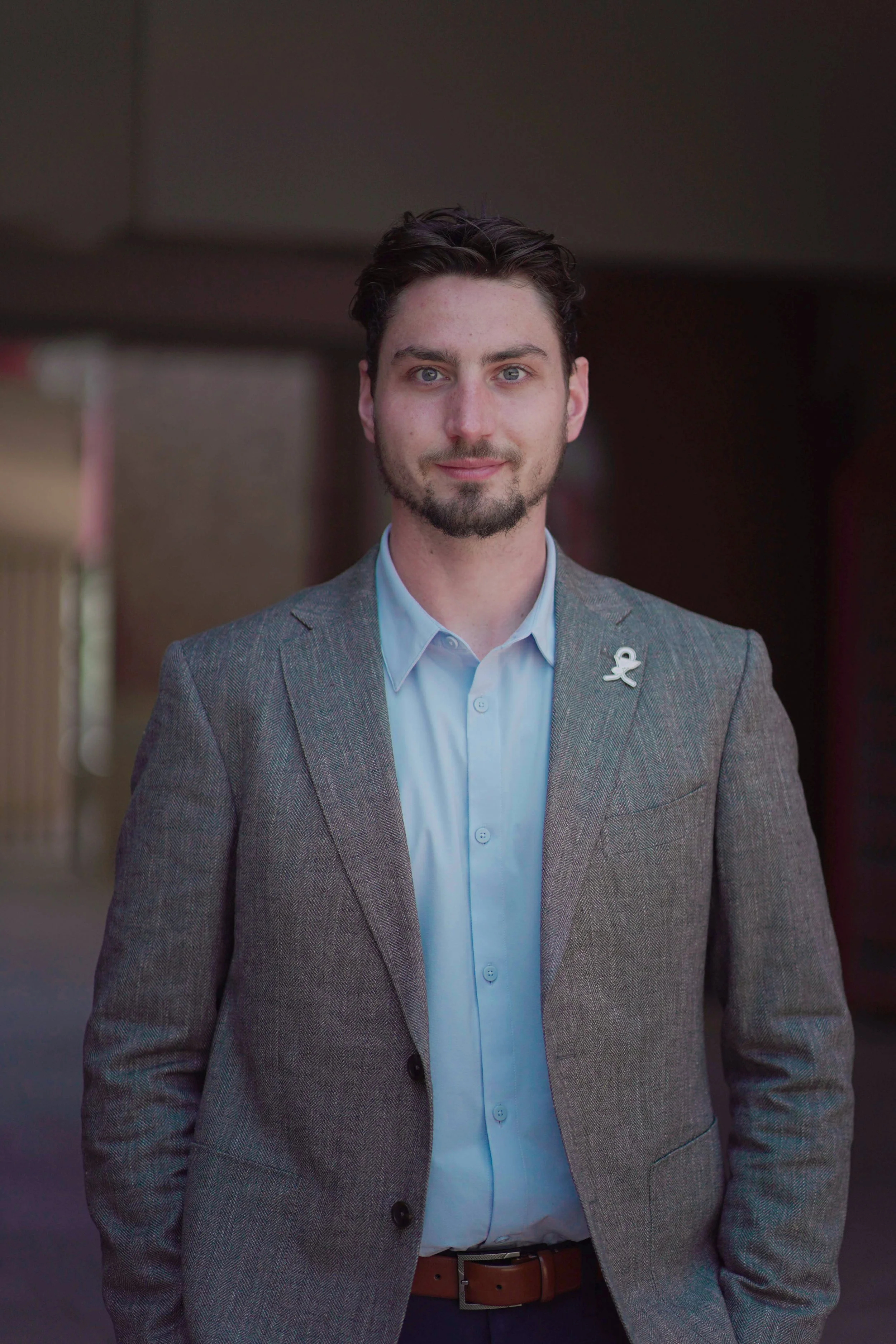 A man with dark hair, light skin, and blue eyes, wearing a light blue dress shirt, gray blazer with a white lapel pin, and a brown belt, standing indoors with a dark background.