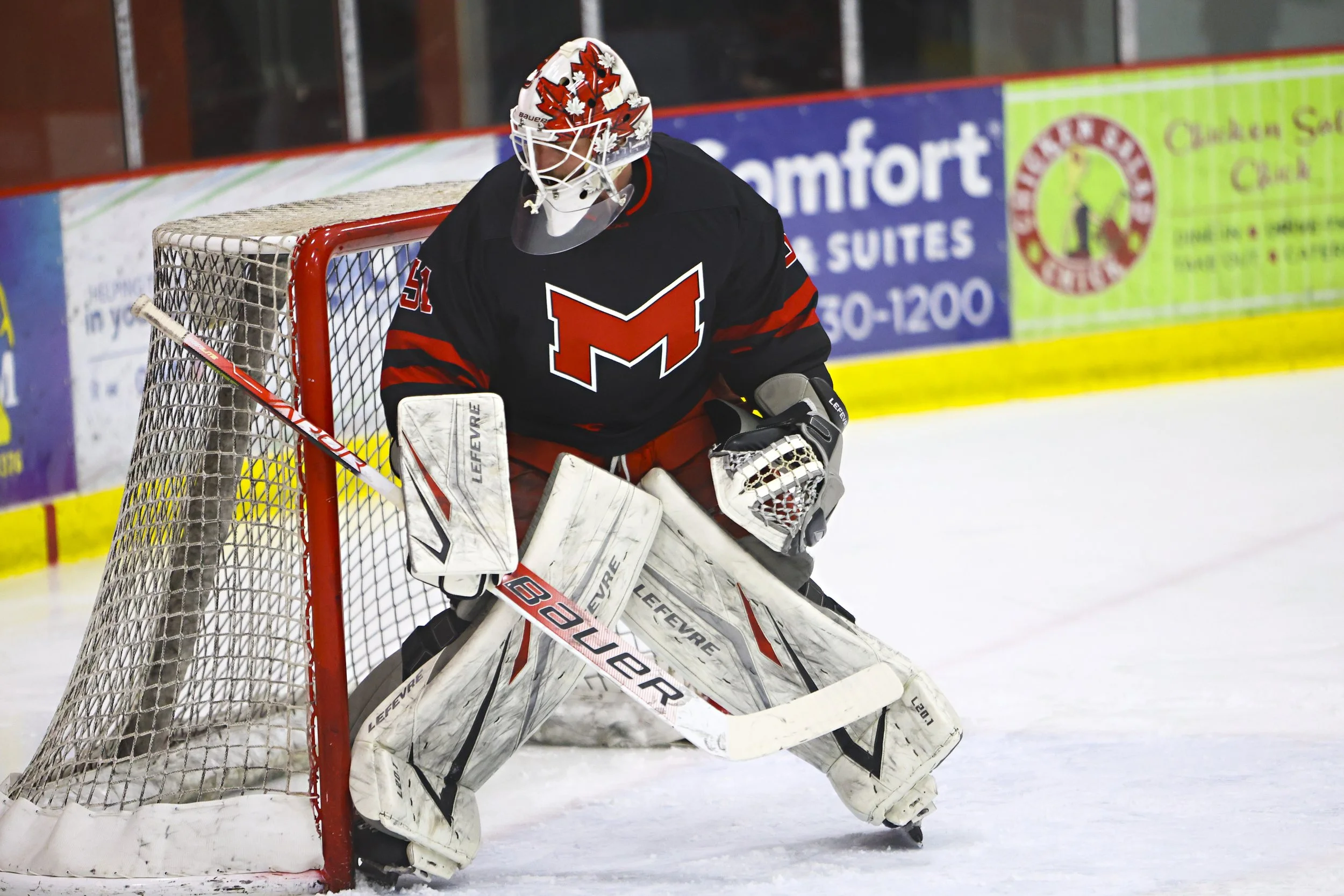 An ice hockey goalie in full gear stands near the goalpost on an ice rink, wearing a black and red jersey with a large red 'M' logo, a protective mask decorated with a red maple leaf, and white leg pads and gloves, preparing to block a shot.