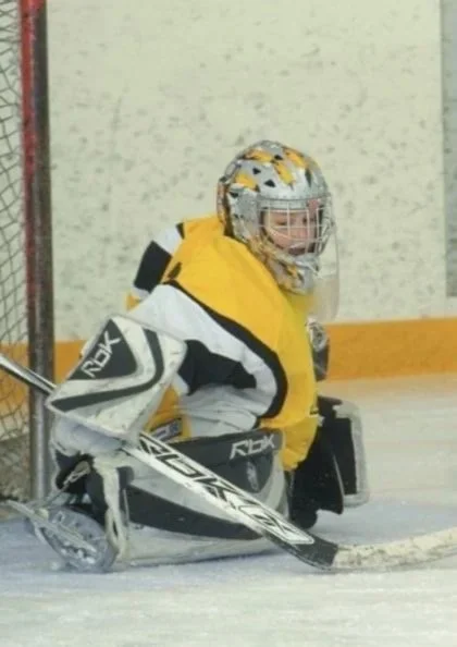 Young ice hockey goalie wearing a helmet, jersey, and pads crouched in front of the net on the ice rink.
