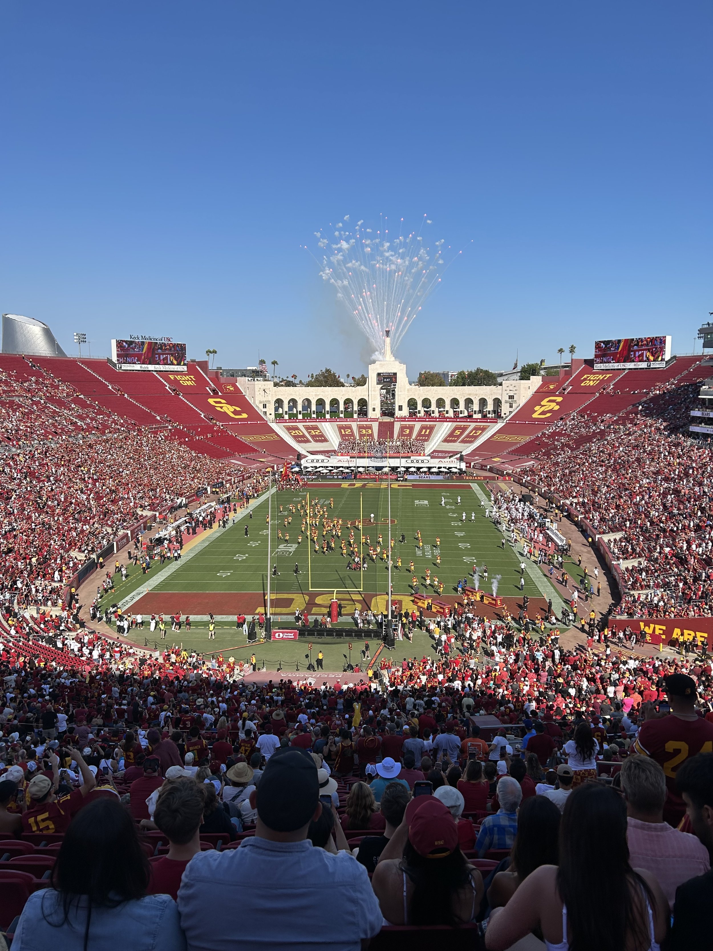 A football stadium filled with fans, with fireworks exploding in the sky above a monument dedicated to USC on game day, during daytime.
