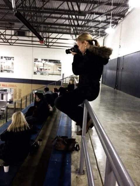 A woman sitting on a rail taking a photo with a camera in an indoor sports arena. Other spectators are seated in the bleachers behind her.