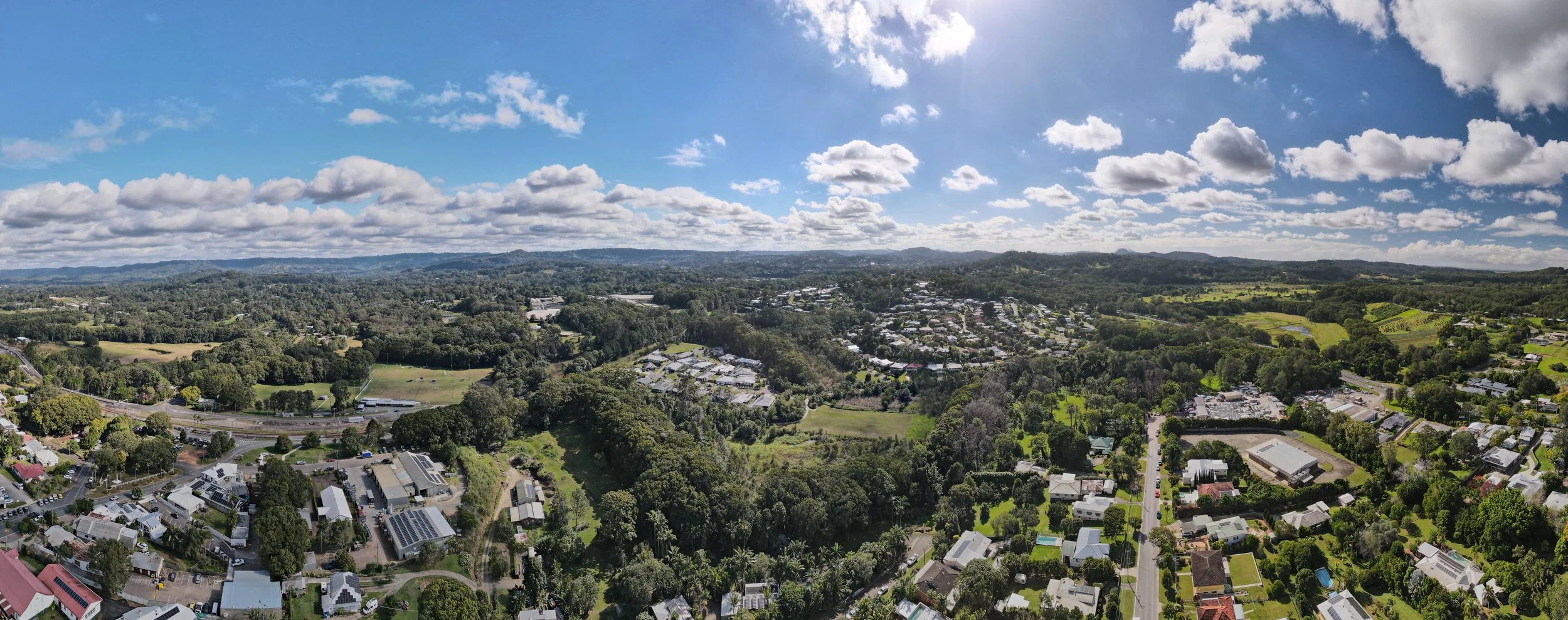 Aerial view of a suburban landscape with houses, trees, open fields, and a blue sky with clouds.