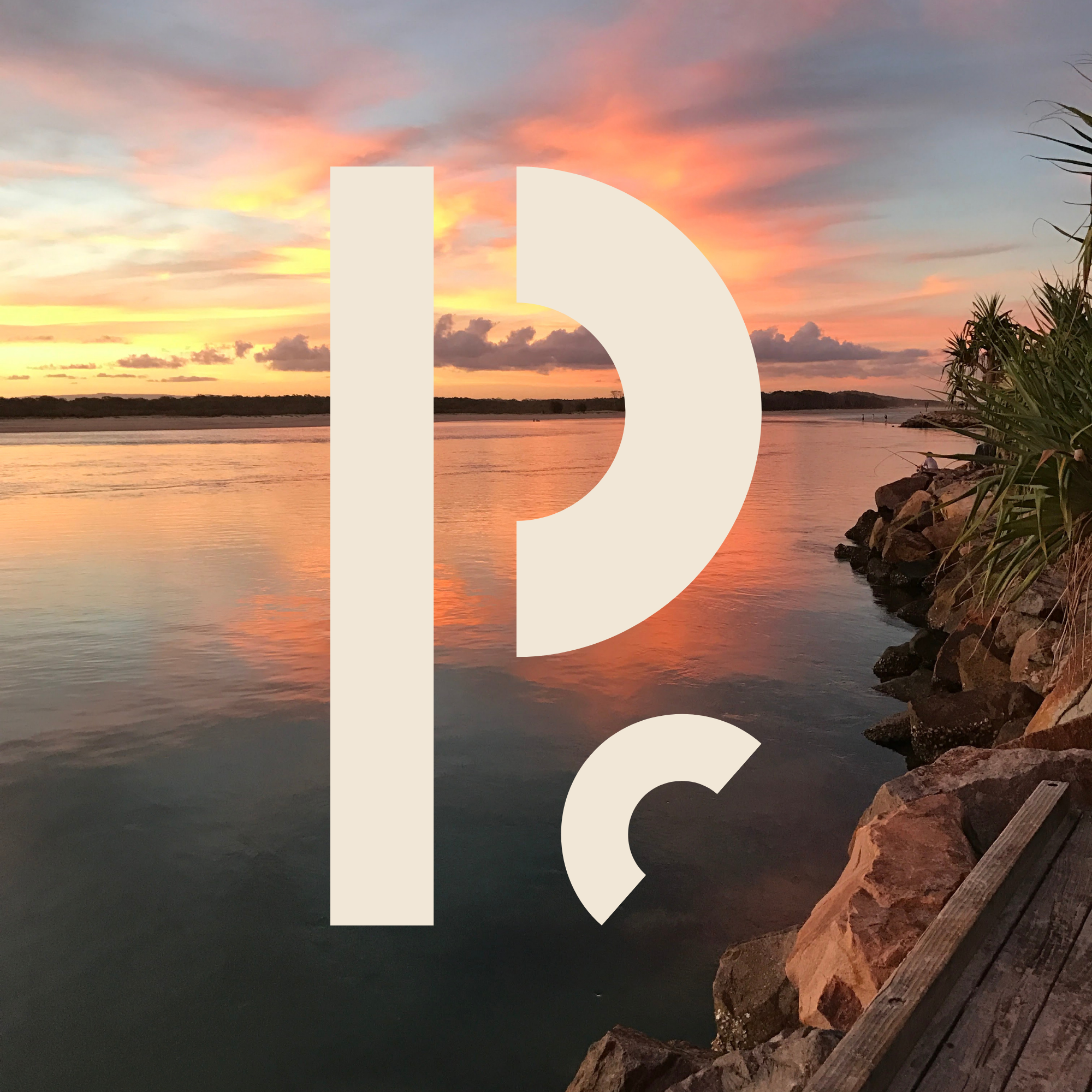 Sunset over a river with a colorful sky, consisting of orange, pink, and purple hues, with a shoreline lined with rocks and some plants.