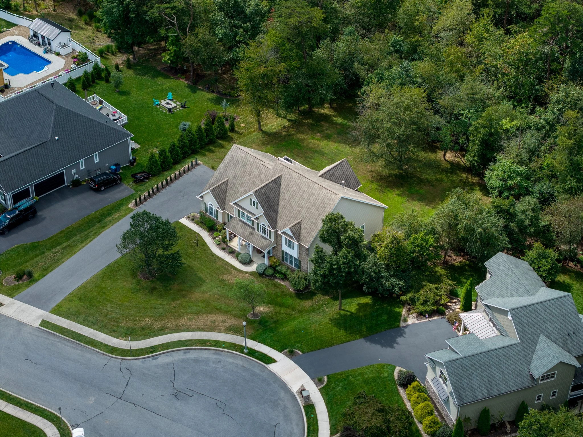 Drone Aerial view of a residential neighborhood with two houses, green yard, trees, driveway, and parked cars, alongside a wooded area. Carlisle, PA
