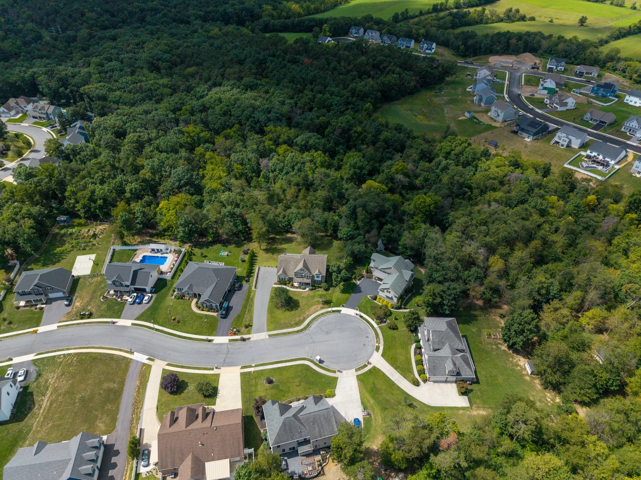 Drone aerial view of a suburban neighborhood with houses, green lawns, and a dense forested area in the center. Carlisle, PA