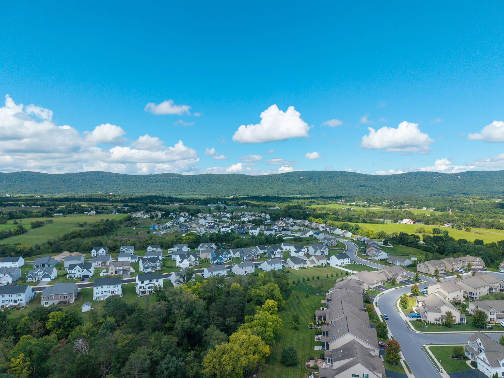 Aerial view of a suburban neighborhood with many houses, surrounded by green trees, with rolling hills and a bright blue sky with scattered white clouds in the background.