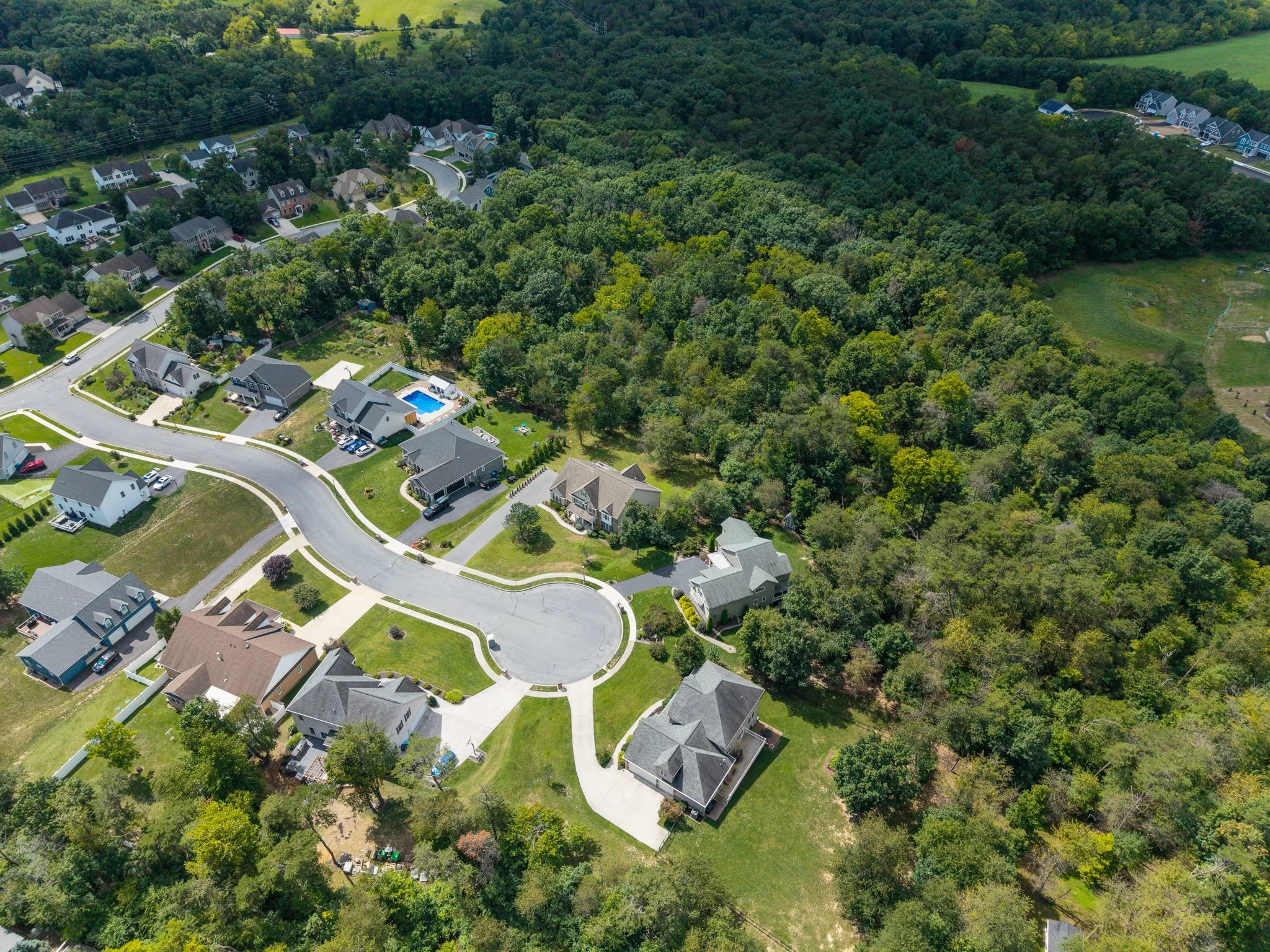 Drone aerial view of a residential neighborhood with houses, roads, and green trees, surrounded by a dense forest. Carlisle, PA