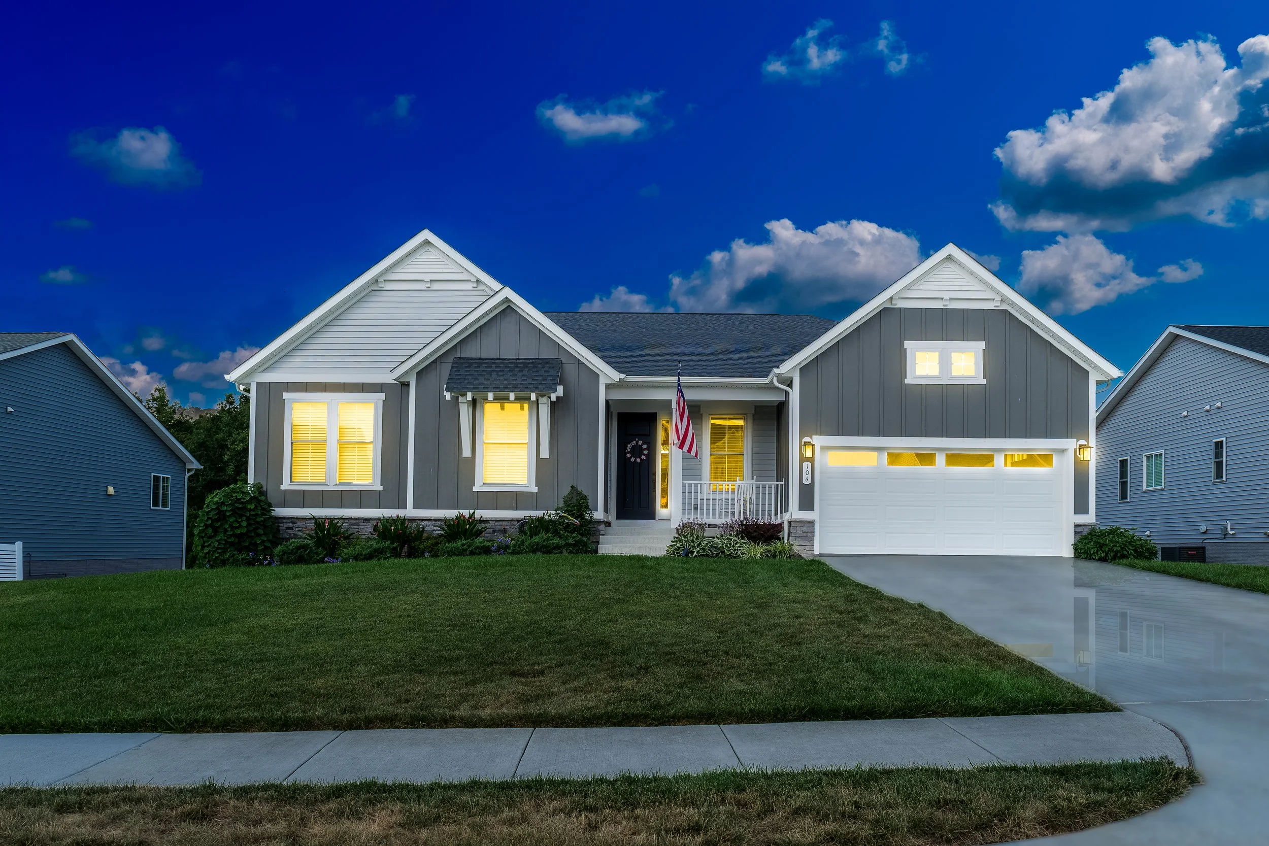 A modern two-story house with gray siding, illuminated windows, and a white garage door, set against a blue sky with scattered clouds during twilight