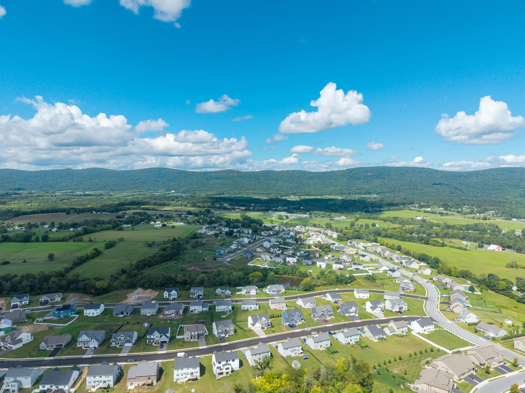 Aerial view of a suburban neighborhood with houses and a winding road, surrounded by green fields and rolling hills, under a partly cloudy blue sky.
