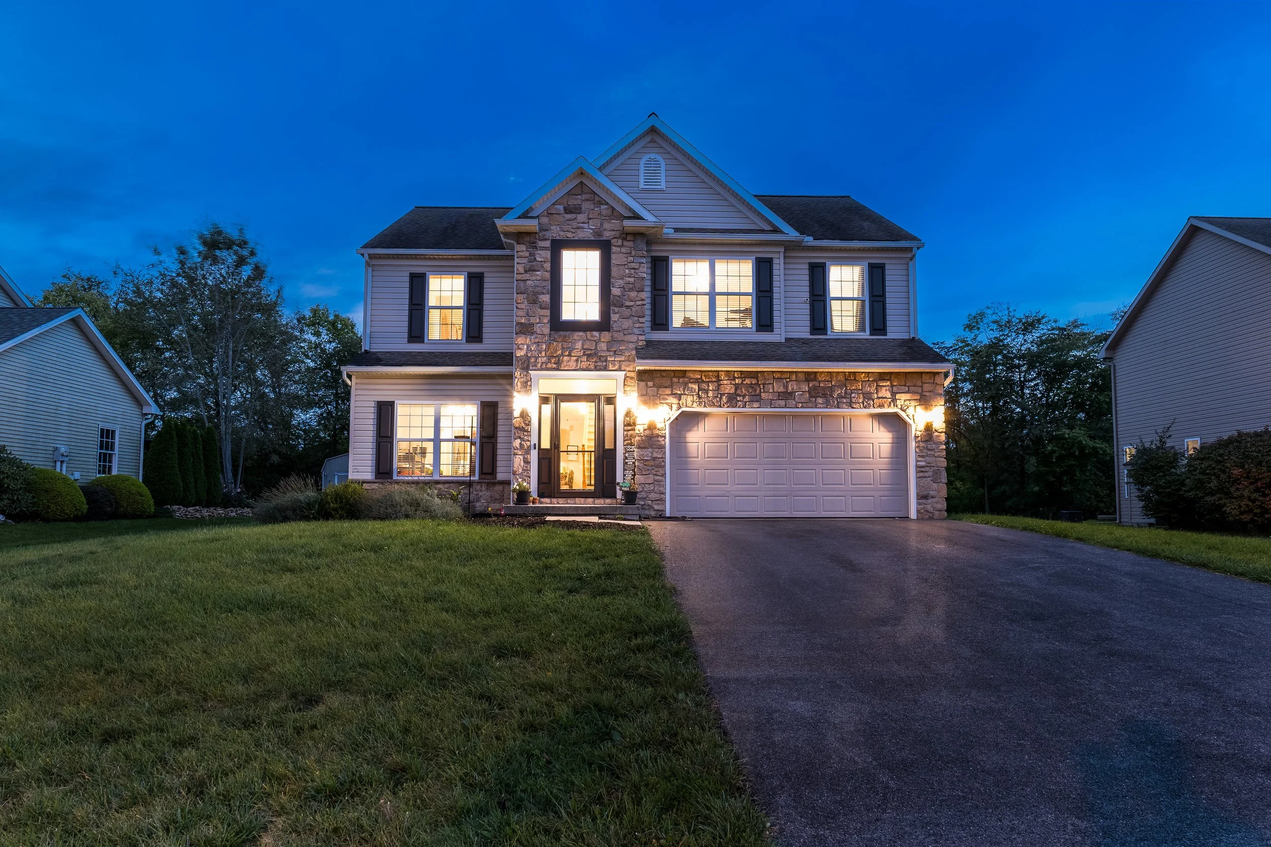 A two-story house with stone and siding exterior, illuminated windows, a paved driveway, and a well-kept lawn during dusk.