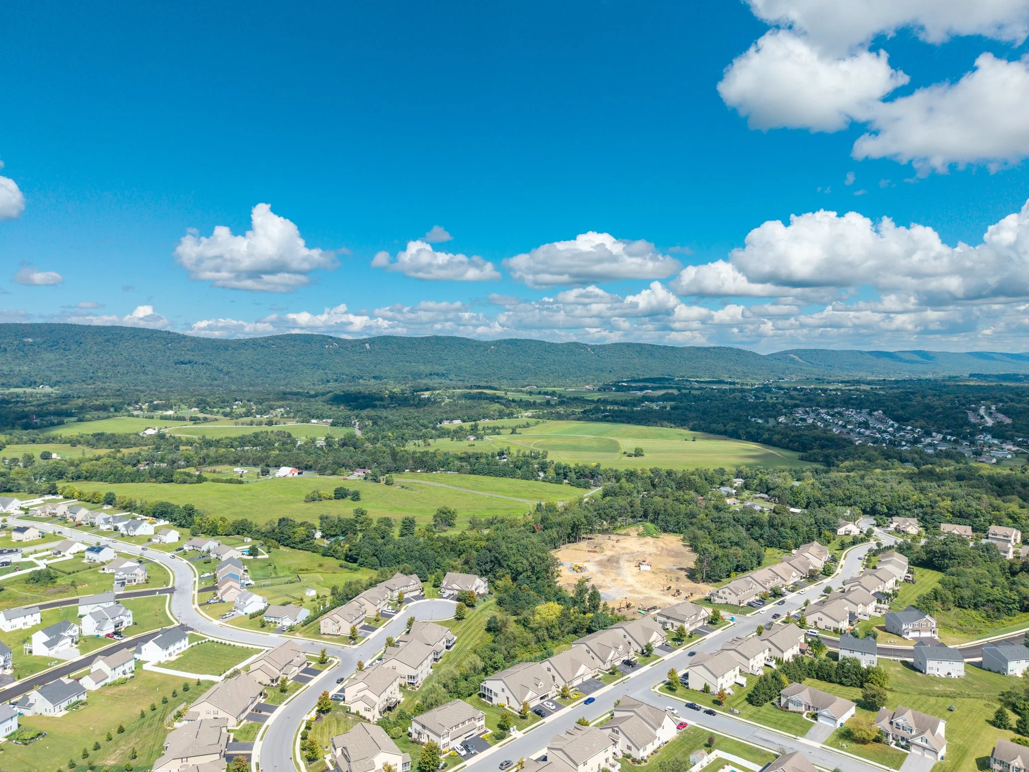 Aerial view of a suburban neighborhood with houses, streets, green lawns, trees, rolling hills, and a partly cloudy blue sky.