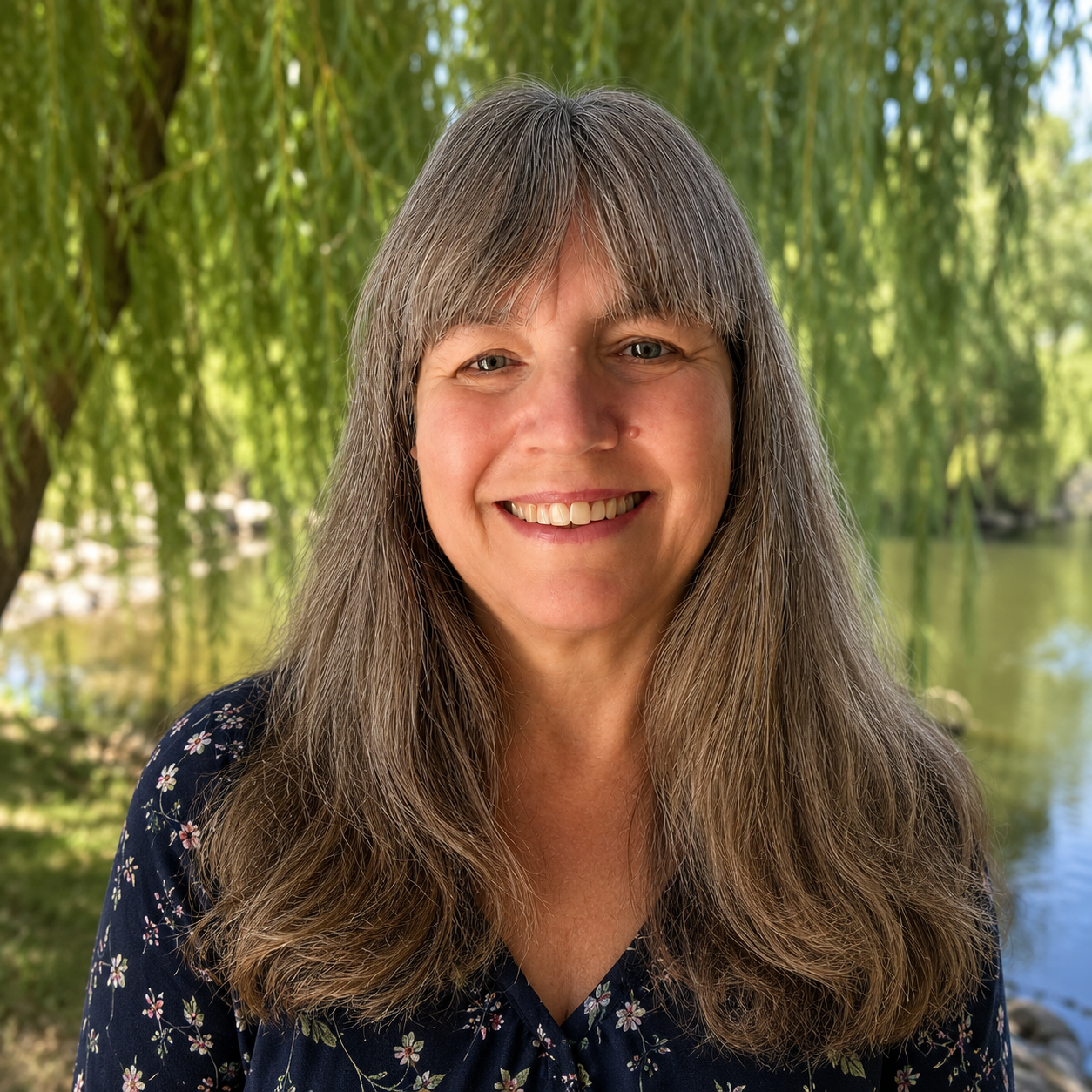 Smiling woman with long gray hair outdoors near a body of water and weeping willow trees in the background.