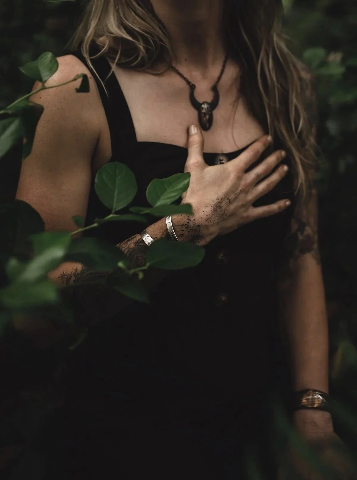 A woman with long hair is wearing a black sleeveless top, a skull necklace, and jewelry. She has tattoos on her arms and hands and is standing amidst green foliage. She is holding her chest with one hand, and her face is not visible.