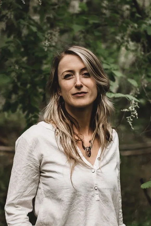 A woman with light brown and blonde hair standing outdoors in front of green foliage, wearing a white button-up shirt.