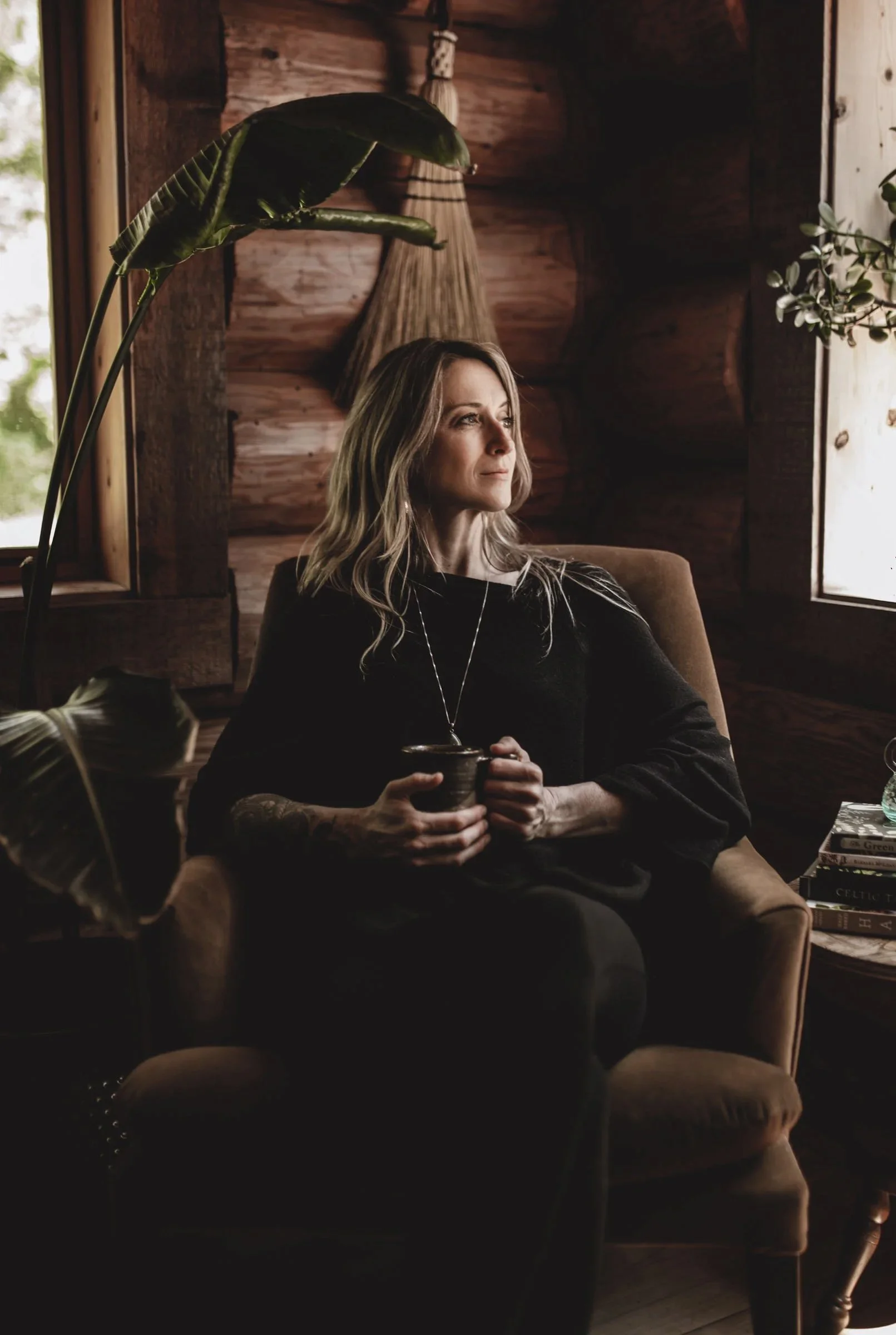 A woman with blonde hair sitting in a cozy wooden room, holding a mug, looking thoughtfully out the window, with books stacked on a table beside her