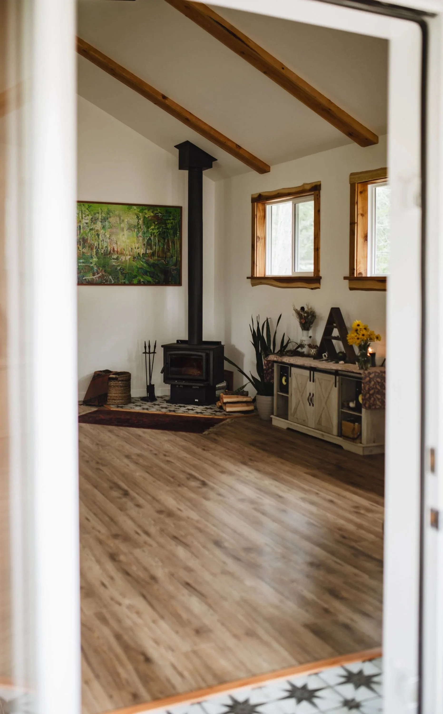 Studio with wood floors, a wood stove, a colorful painting, a console table decorated with flowers and candles, and two windows with wooden frames.
