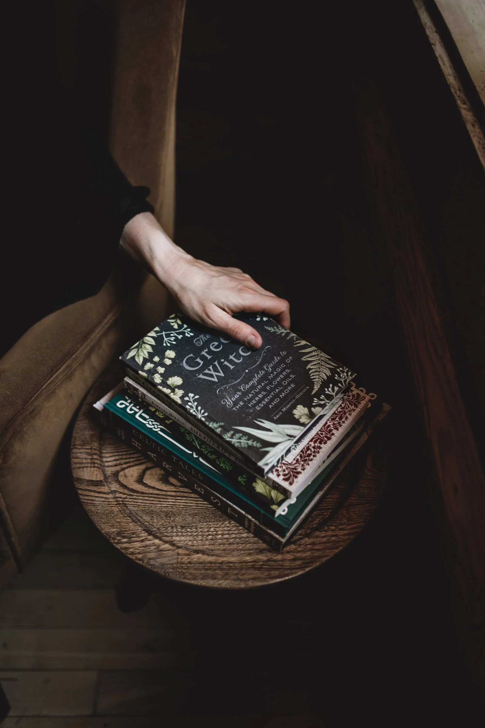 A hand reaching for a book titled 'The Green Witch' on a stack of books on a round wooden table in a dimly lit space.