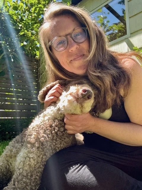 A woman with long brown hair and glasses taking a selfie outdoors with her curly-haired dog, sitting in a garden with green foliage and a house in the background.
