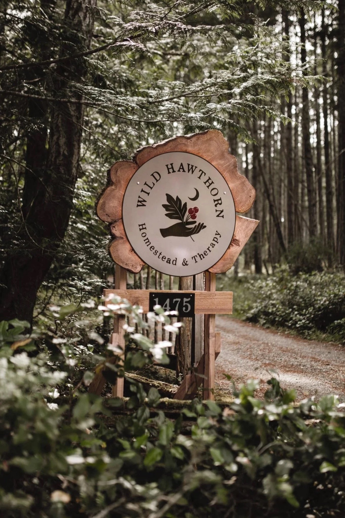 A wooded trail in a forest with a sign that reads 'Wild Hawthorn - Homestead & Therapy' and a smaller sign below with the number '1475', surrounded by green foliage and tall trees.