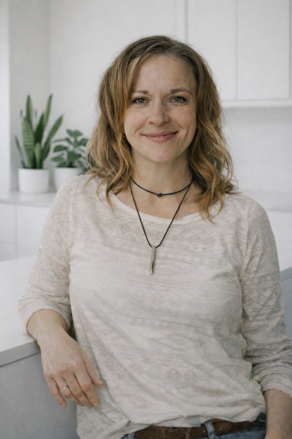 A young woman with shoulder-length wavy red hair smiling at the camera, wearing a light-colored patterned long-sleeve shirt and layered necklaces, standing in a white kitchen with green potted plants in the background.