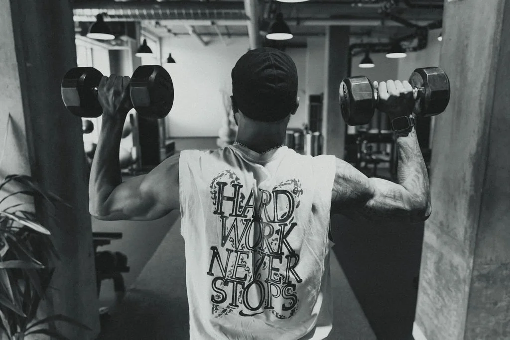Man lifting dumbbells in a gym, wearing a sleeveless shirt with the text "Hard Work Never Stops" on the back.