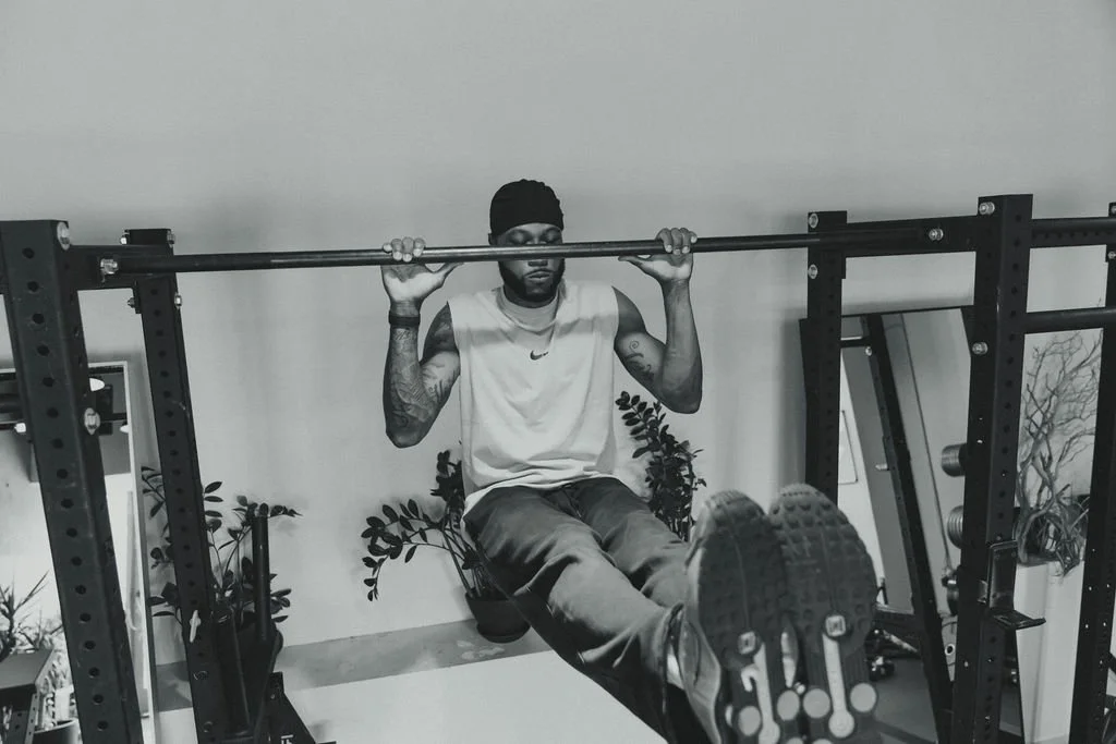 A man in a sleeveless shirt and beanie doing leg raises on a gym bench, holding a horizontal bar.
