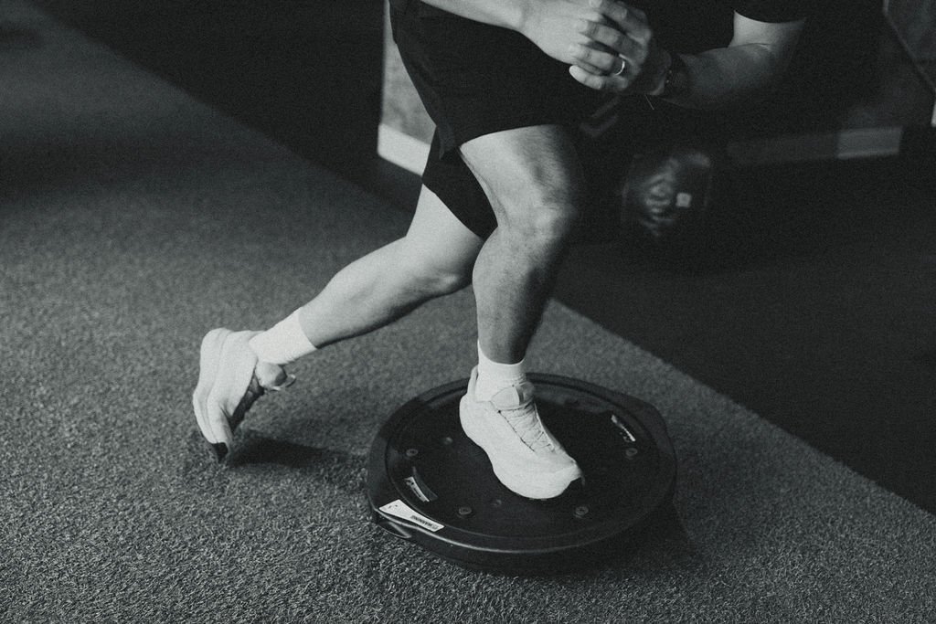 Person standing on a balance board in a gym, wearing athletic shorts, a t-shirt, and sneakers.