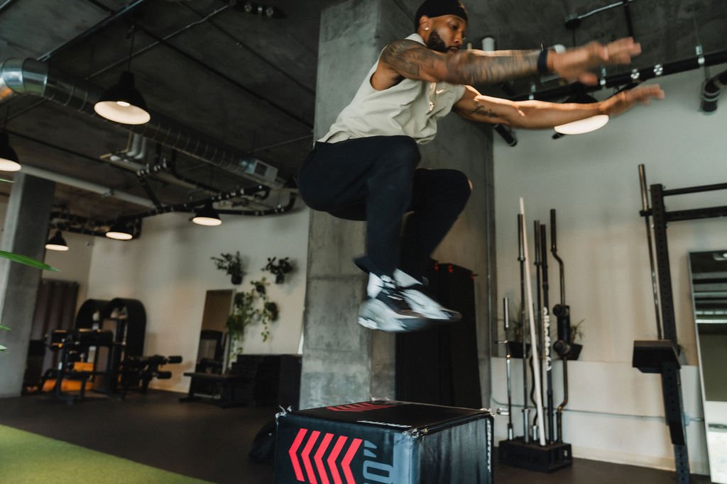 A man in athletic clothing performing a jump onto a plyometric box in a gym.