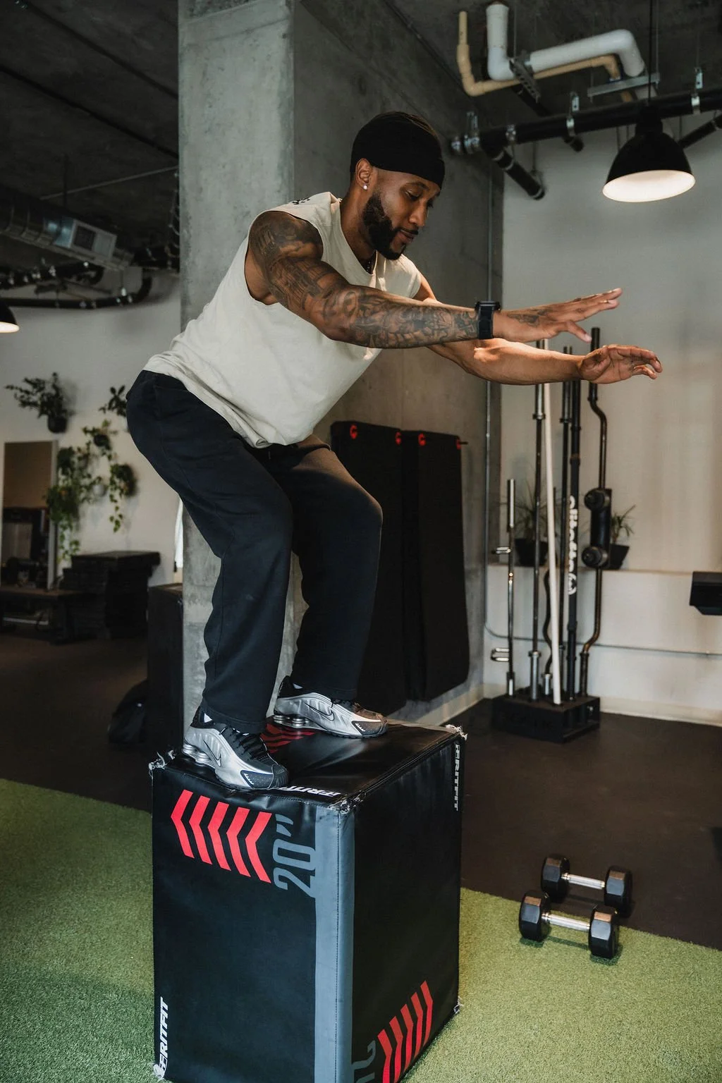 A man wearing a black headband, white sleeveless shirt, black pants, and sneakers is balancing on a large black plyometric box during a workout in a gym. His arms are extended outward for balance, and he appears to be preparing for a jump.