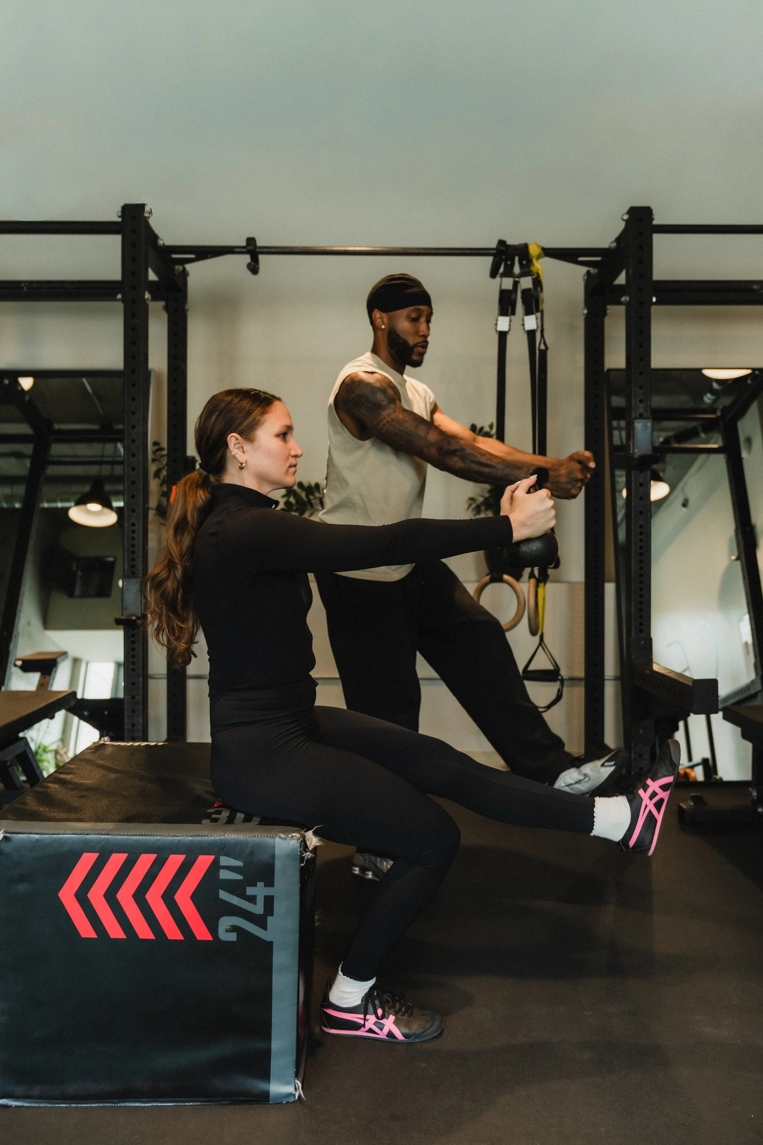 A woman sitting on a workout mat performing a sit-up exercise, with a man standing behind her holding a kettlebell, in a gym setting.